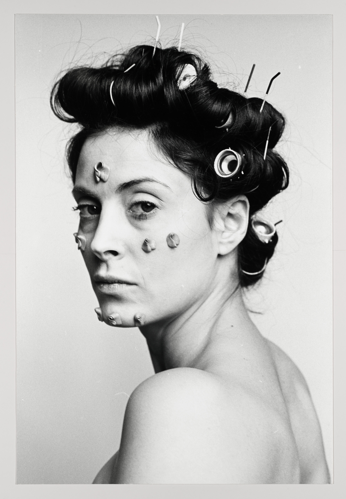 A black and white portrait of a woman with an artistic hairstyle featuring hair rollers and bobby pins. She has several snails placed on her face and neck, creating a surreal effect. The woman is looking off to the side with a neutral expression, and her shoulders are bare.