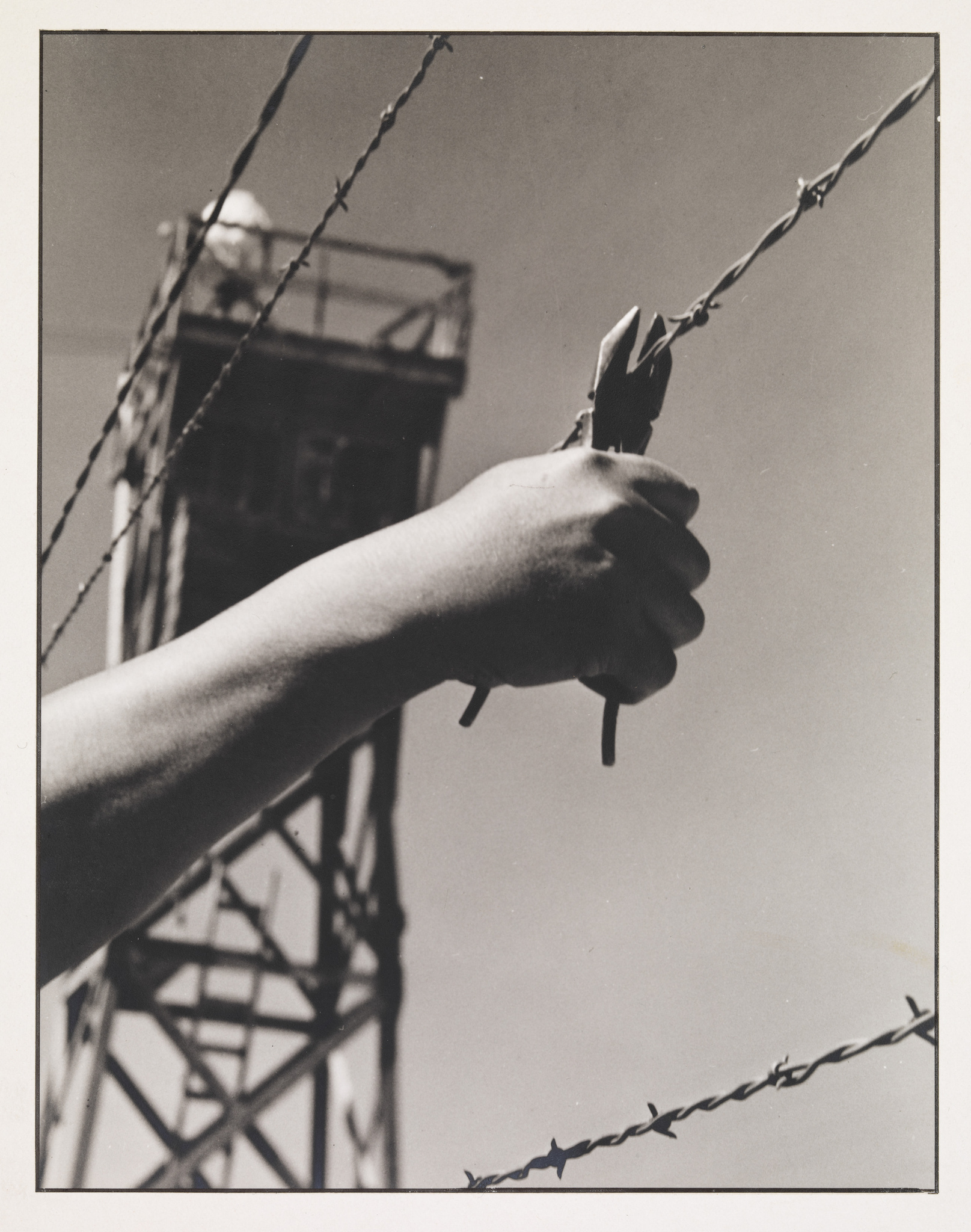 A hand holding wire cutters near barbed wire, with a watchtower in the background against a clear sky.