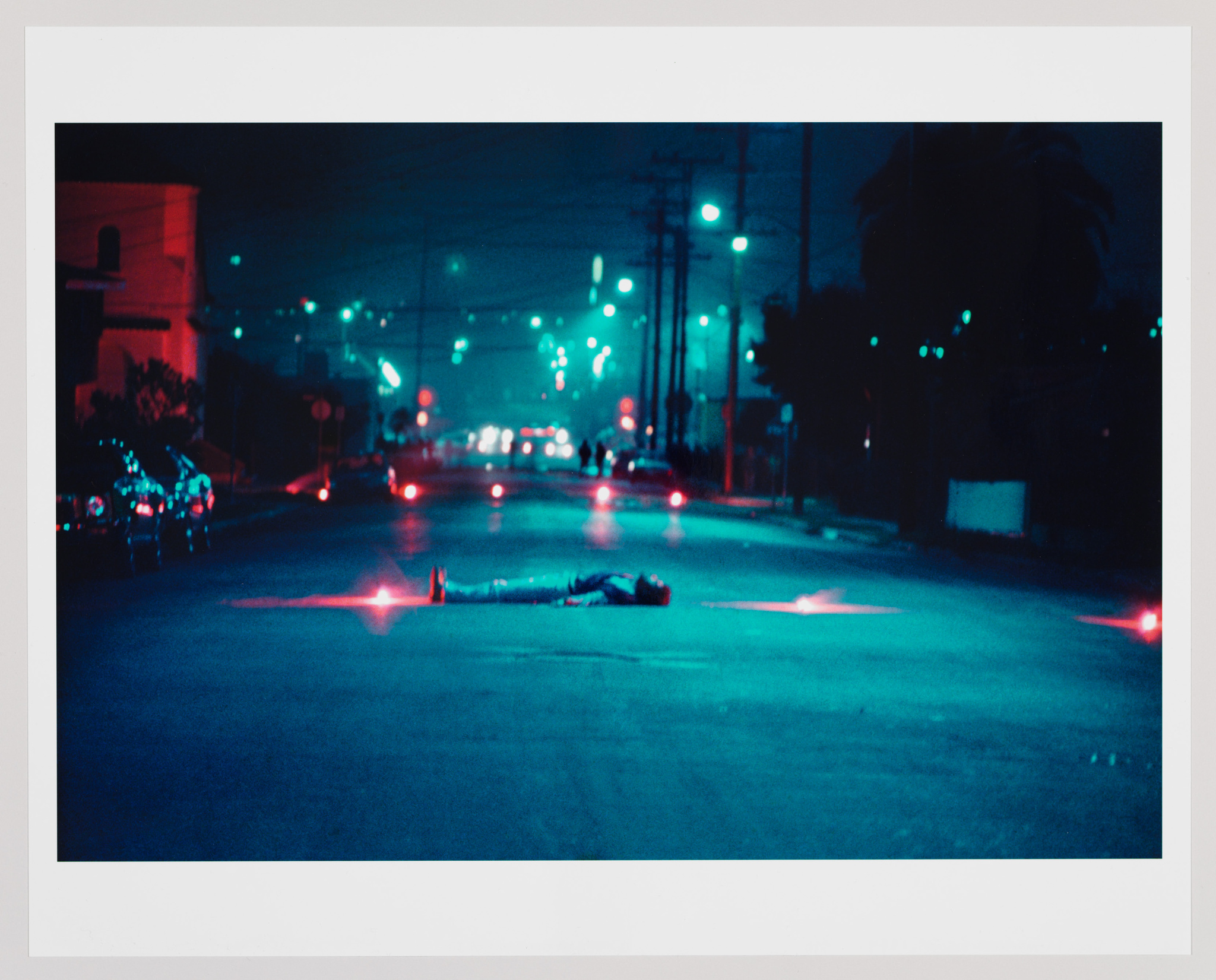 A nighttime street scene with a moody, blue-toned atmosphere. Streetlights and traffic signals cast a glow on the pavement, and red flares are positioned around a figure lying in the road, suggesting an emergency or crime scene. The environment is urban with buildings lining the street, and the focus is on the illuminated, solitary figure on the ground.