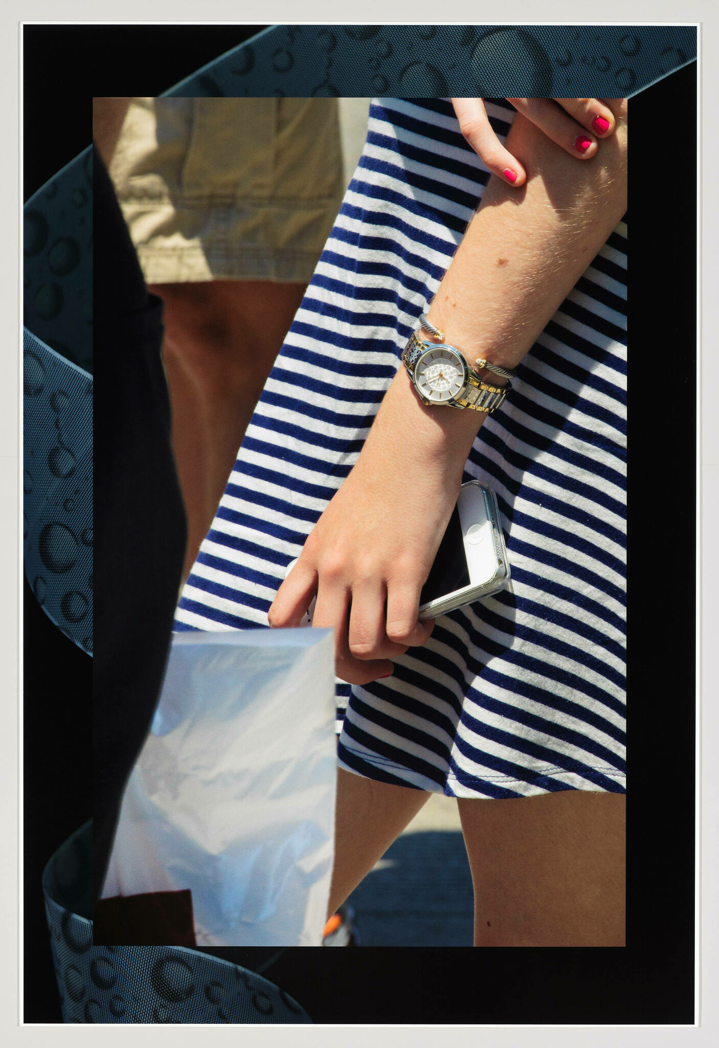 A close-up of a person's arm wearing a gold watch, with a striped blue and white dress and holding a smartphone and a plastic bag, with a partial view of a beige shorts in the background.