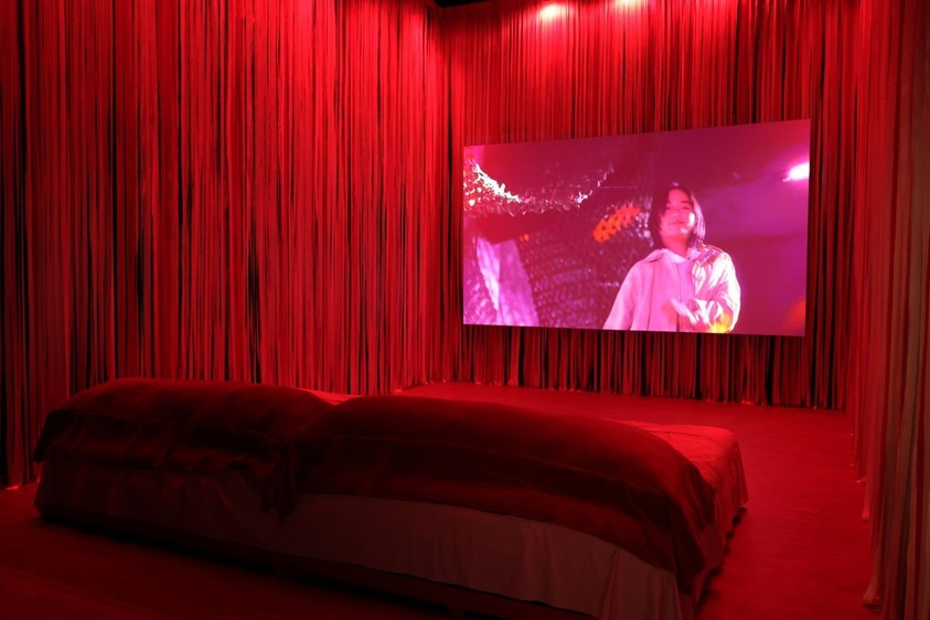 A room bathed in red light features a large bed and a screen displaying a person in a white shirt.