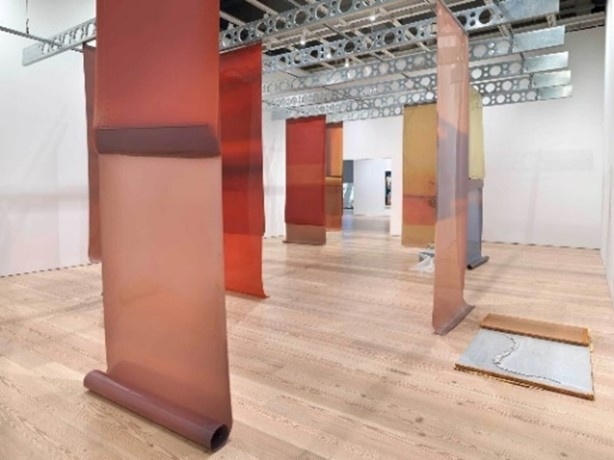 Art installation with large, translucent colored panels hanging in a gallery space with wooden floors and metal ceiling structures.