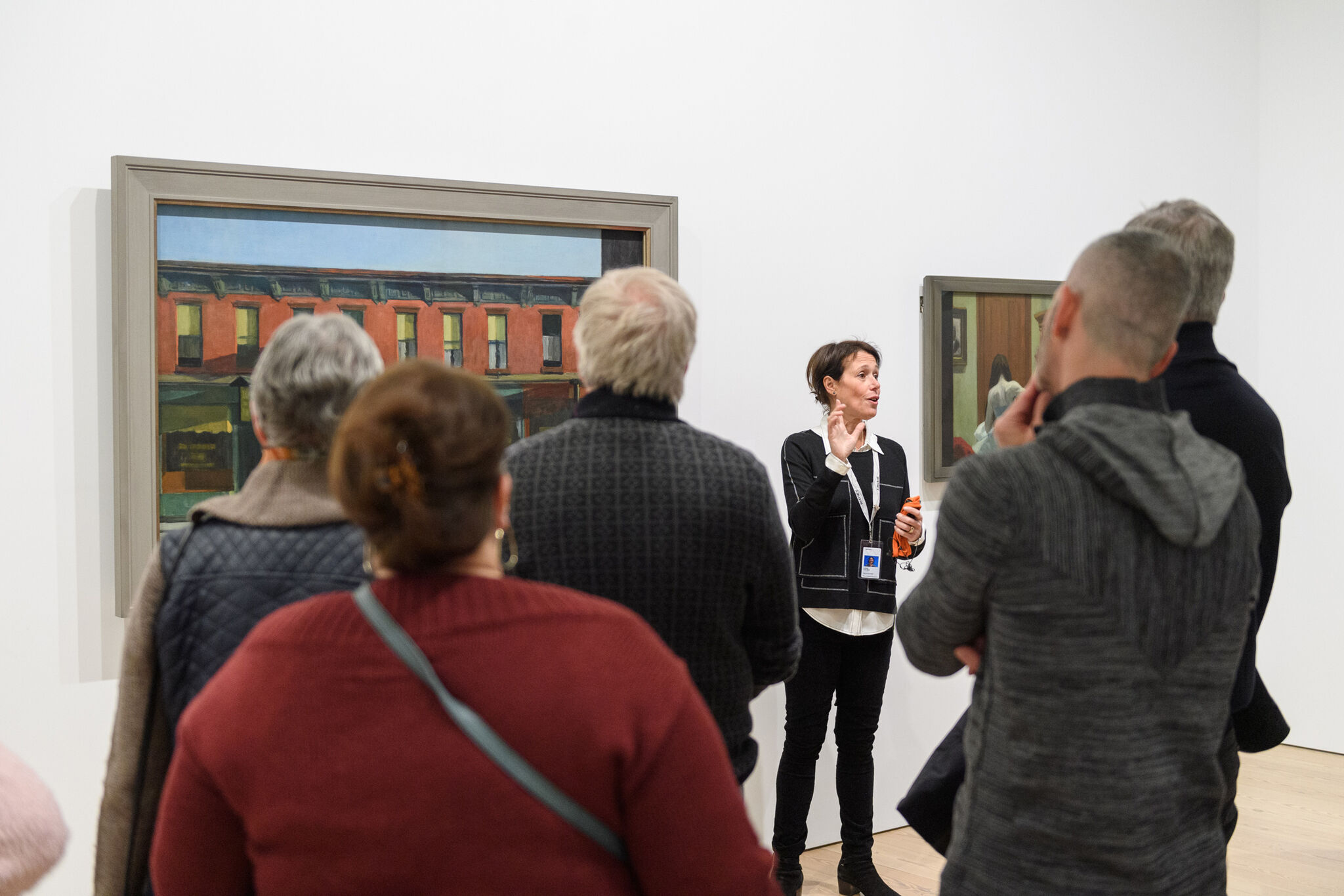 A museum guide speaks to a group of visitors in front of a painting of a red brick building.