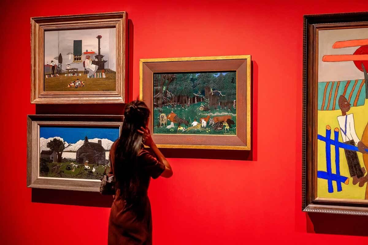 A woman observes colorful paintings on a vibrant red gallery wall, featuring various scenes and styles.