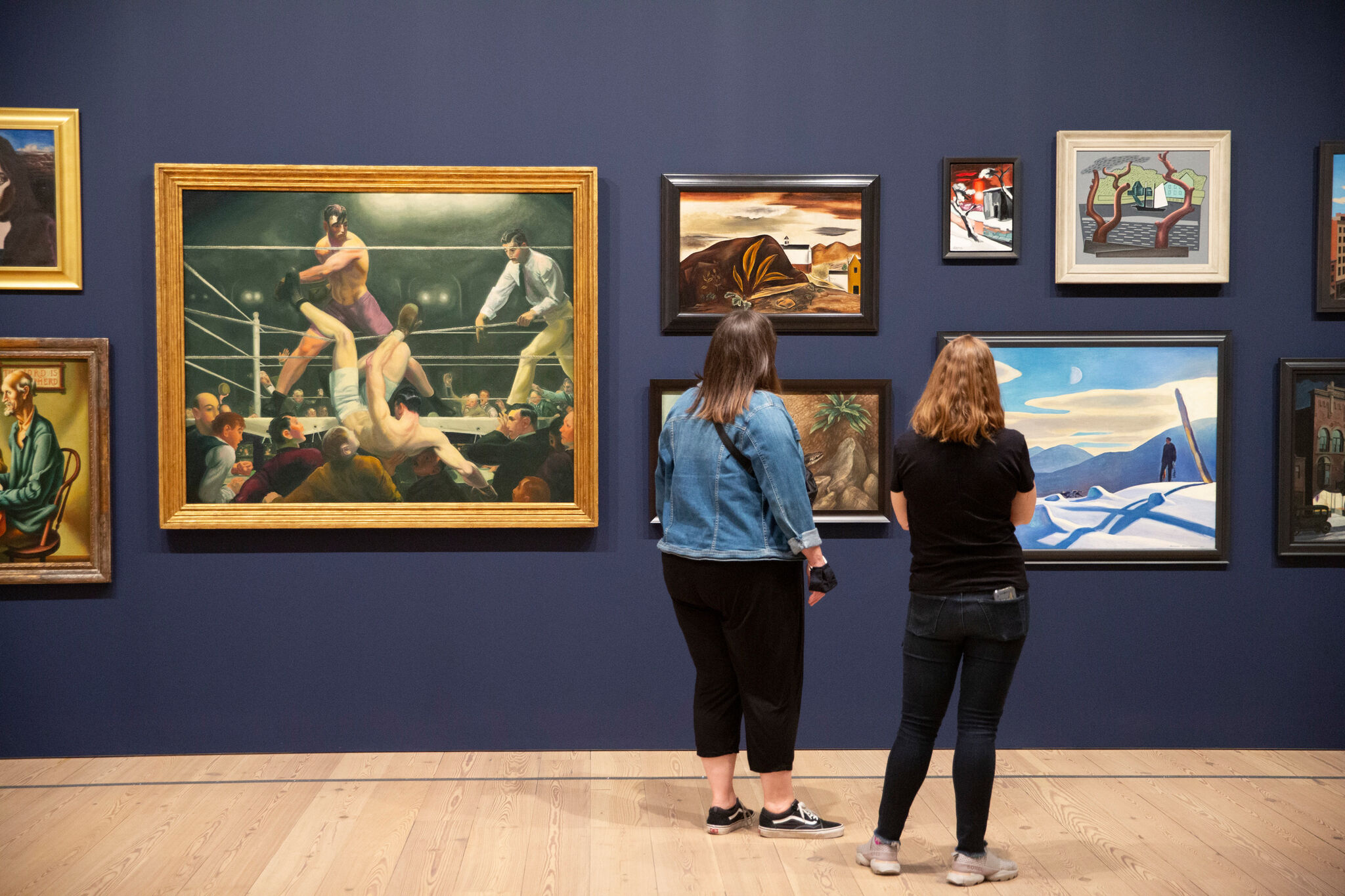 Two people stand in a gallery with blue walls and many paintings. 