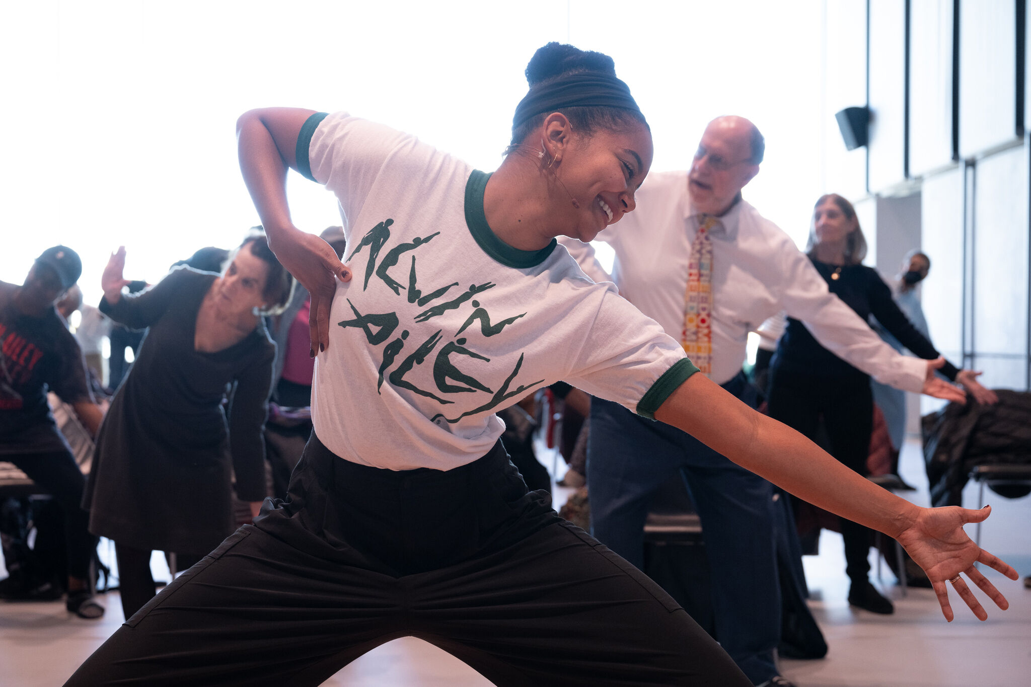 A woman in an "Alvin Ailey" shirt leads a diverse group in a dance class, smiling and extending her arms energetically.