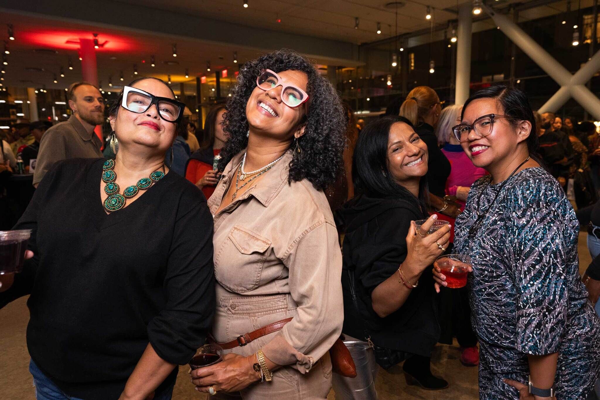 Four women smiling and posing at a lively indoor event, holding drinks, with a crowd and colorful lighting in the background.