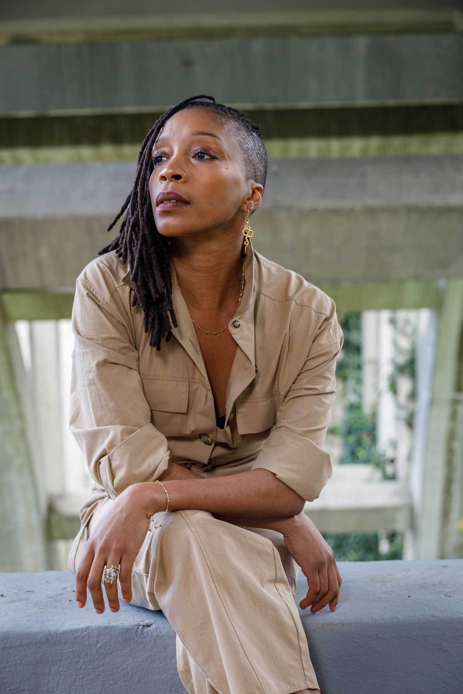 Person with dreadlocks and a side shave, wearing a beige jumpsuit, sits pensively under a concrete structure, adorned with gold jewelry.