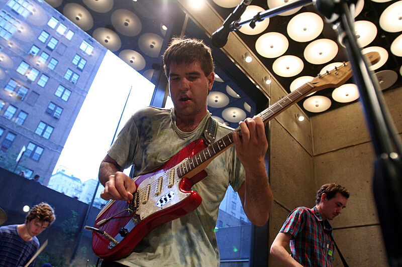 Three musicians perform indoors, with one playing a red electric guitar in the foreground. Large windows and ceiling lights are visible.