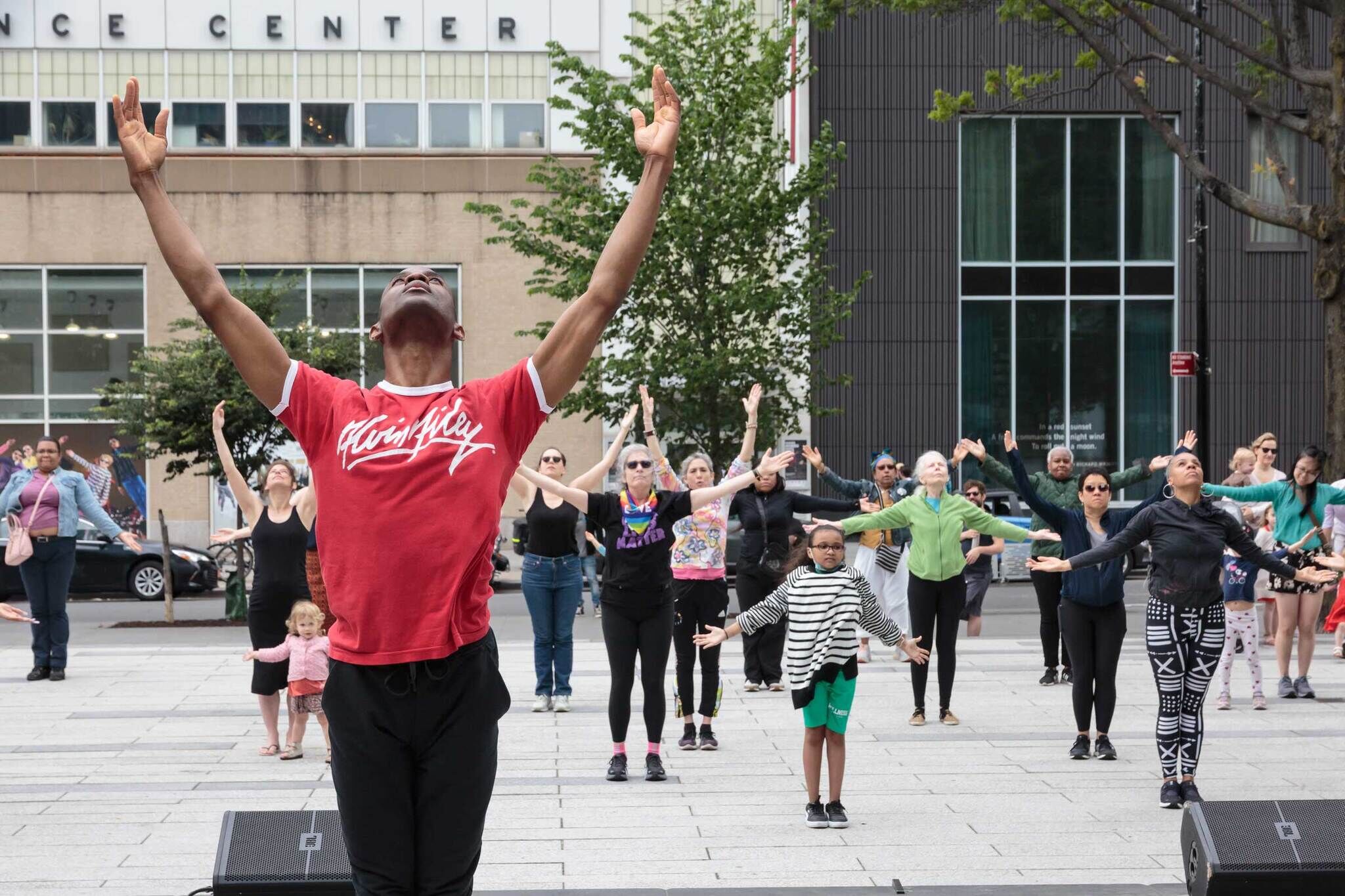 A group of people, led by a man in a red shirt, participate in an outdoor dance or exercise class, raising their arms in unison.