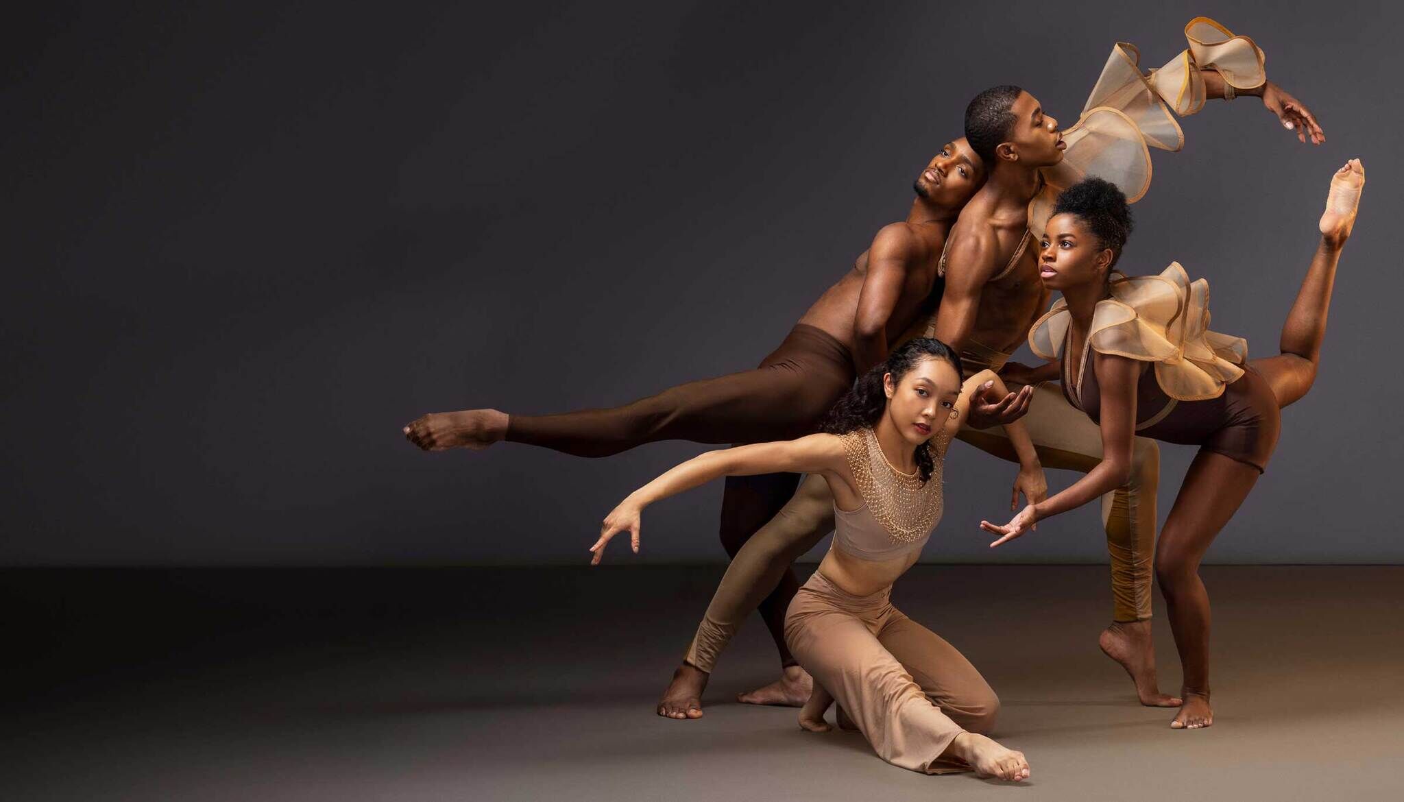 Four dancers in dynamic poses, wearing earth-toned costumes, against a dark background.