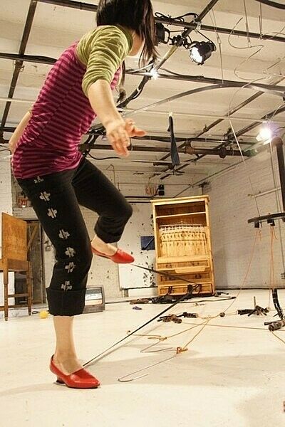 A woman stands on one leg facing away from the camera, with strings and long sticks of metal in front of her.