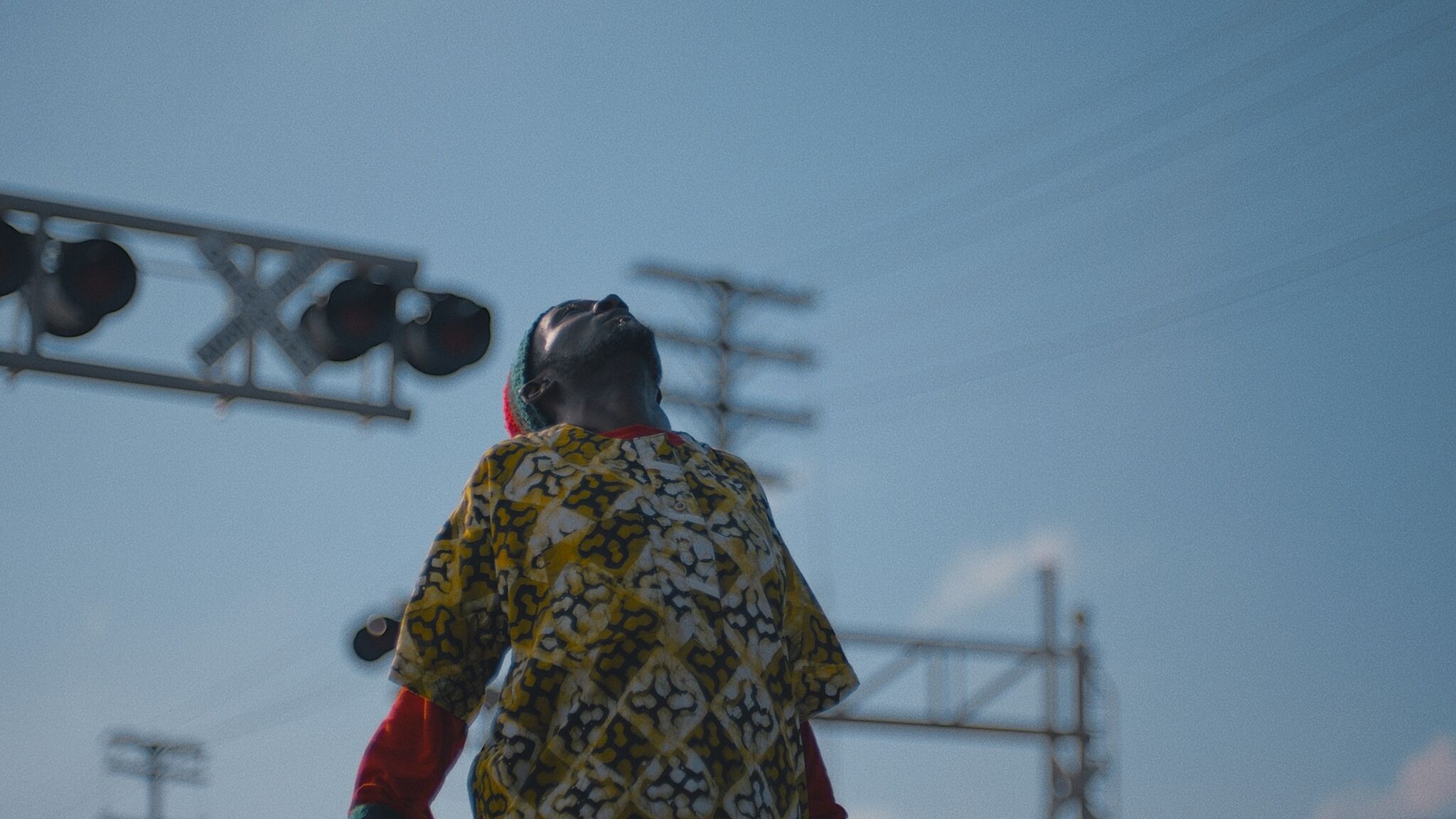 A video still of a figure looking up at the sky in front of a railroad.