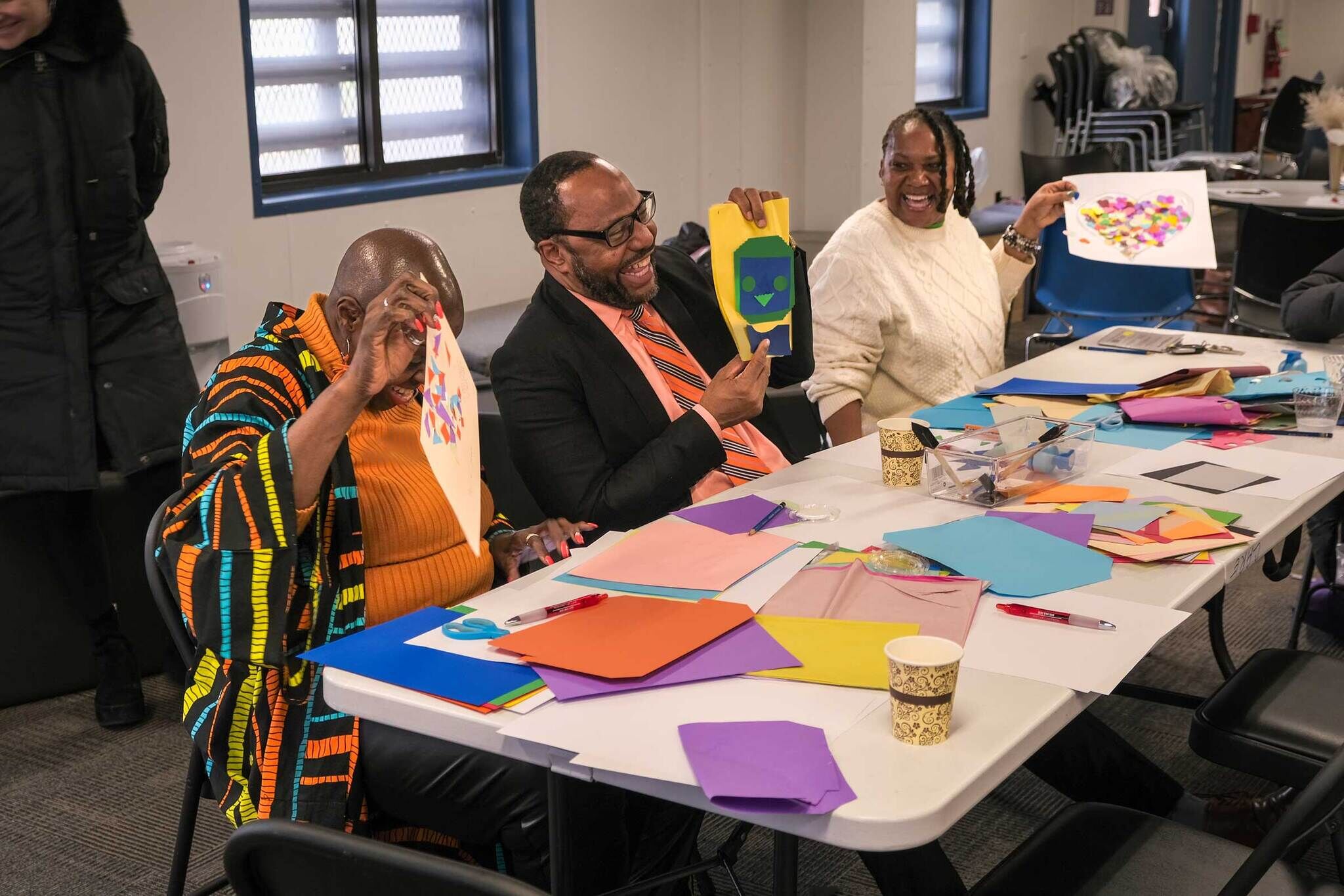 Three people laughing and showing their colorful paper crafts with art supplies scattered on the table.