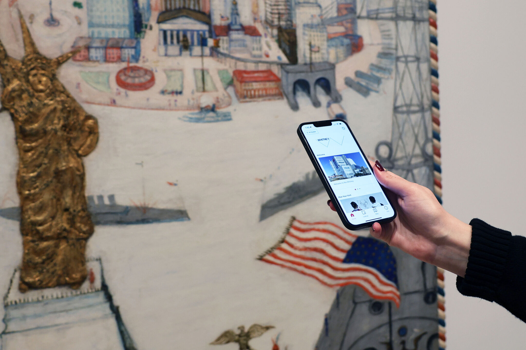 A person's hand holding a smartphone with an image on its screen, in front of a colorful, abstract painting with a statue on the left.