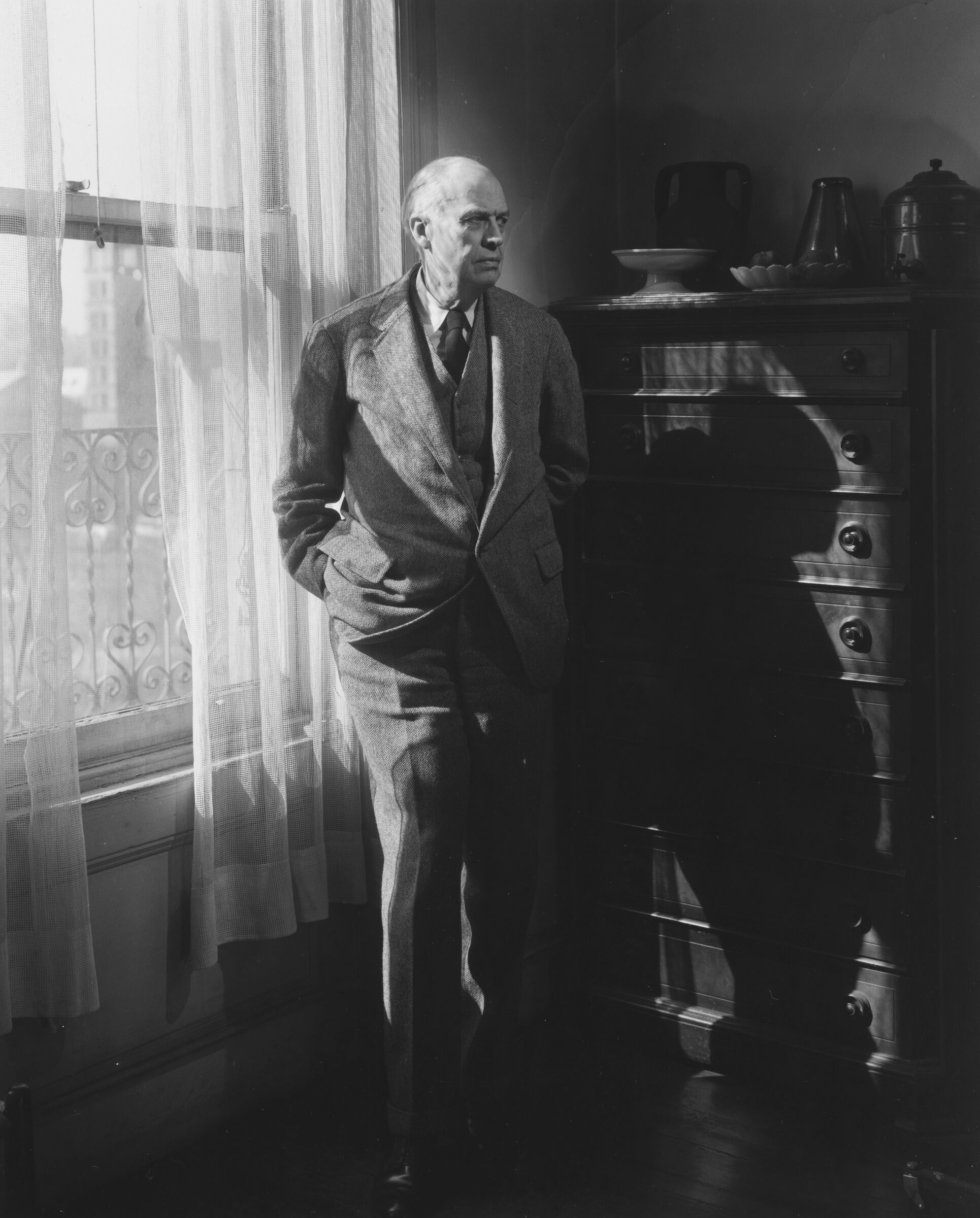 A photograph of Edward Hopper standing by a window and chest of drawers. 