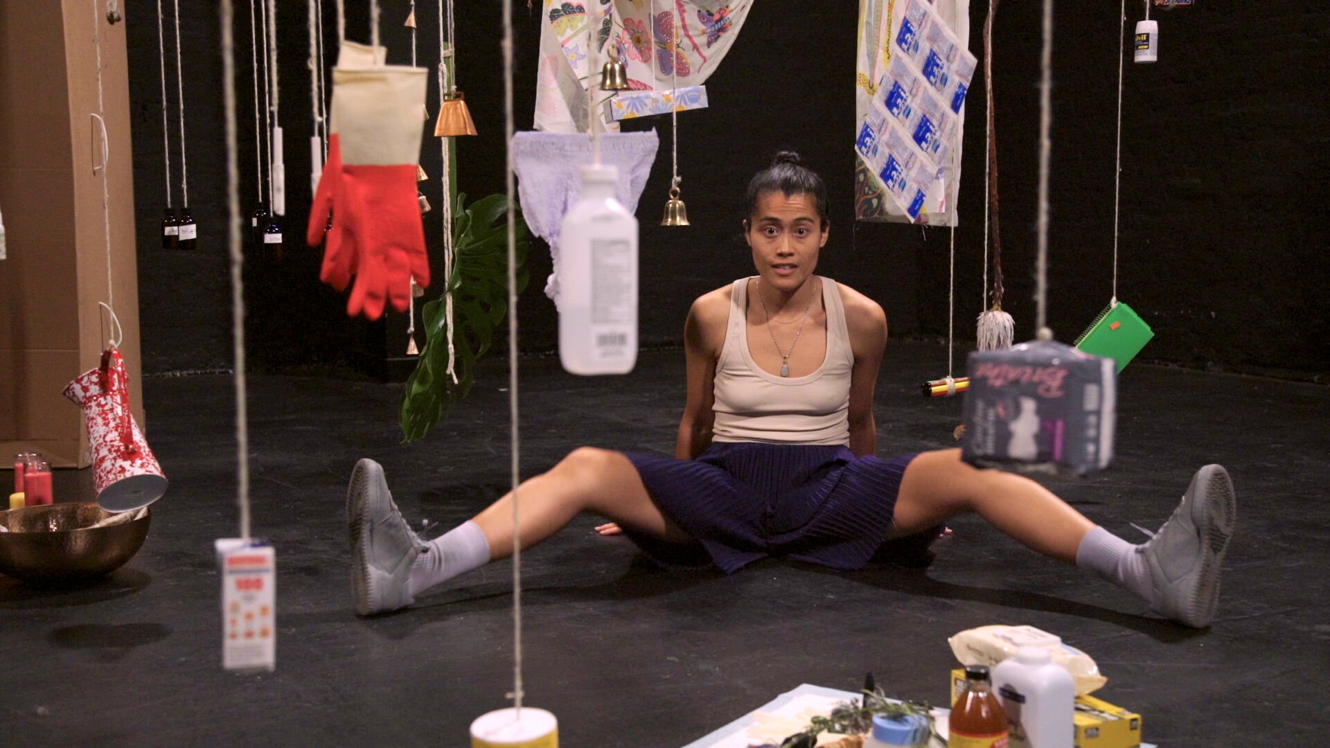 A performer, wearing a tan tank top, a purple pleated skirt, and grey socks and sneakers, sits with her legs spread in a V-shape. She is framed on either side by hanging objects suspended by string: among them, red gloves, bells, a bottle of medical alcohol, bandages, and a bottle of Advil.