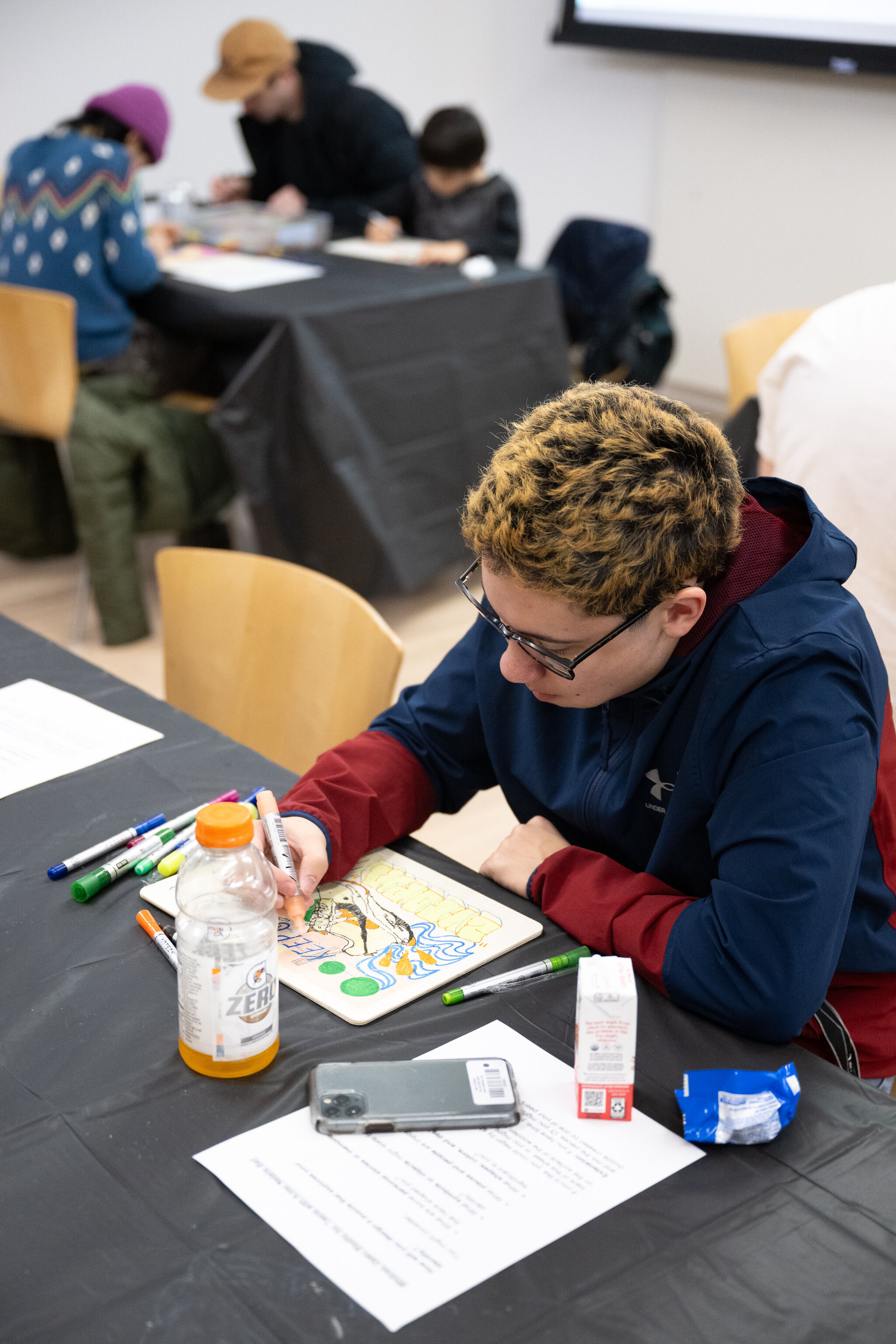 A young person with short, curly blonde and brown hair, wearing glasses and a navy and red jacket, is seated at a table engaged in an art activity. They are using colorful markers to draw on a wooden panel with intricate designs and bold text. The table is covered with a black cloth and has various art supplies scattered around, including markers, a bottle of orange-flavored drink, a small carton of juice, a blue snack wrapper, and a smartphone. In the background, other participants, including a child and two adults, are also engaged in art-making at a separate table. A screen is visible on the wall behind them, indicating an indoor workshop setting.