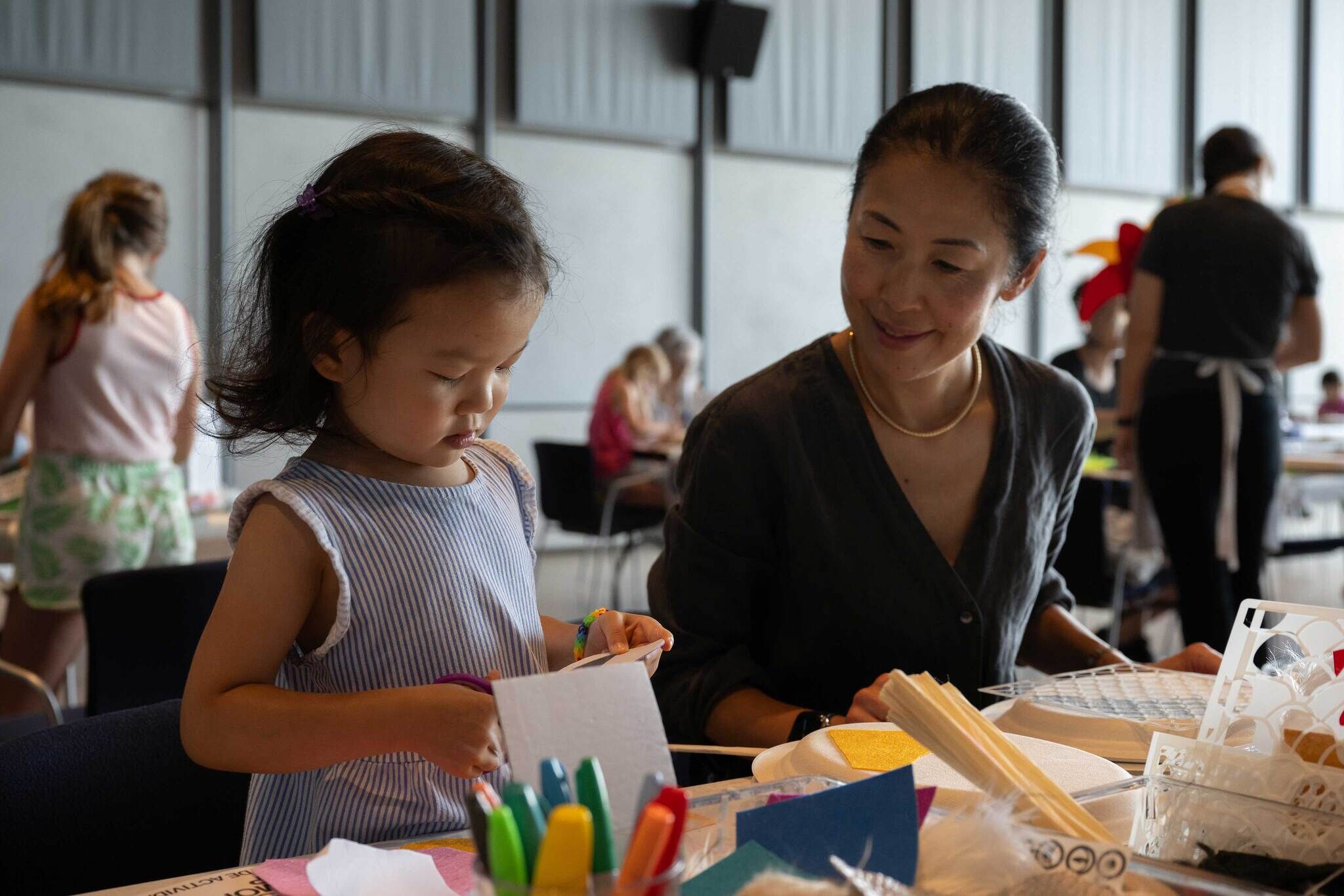 A child cuts a piece of paper at a table while an adult looks on.