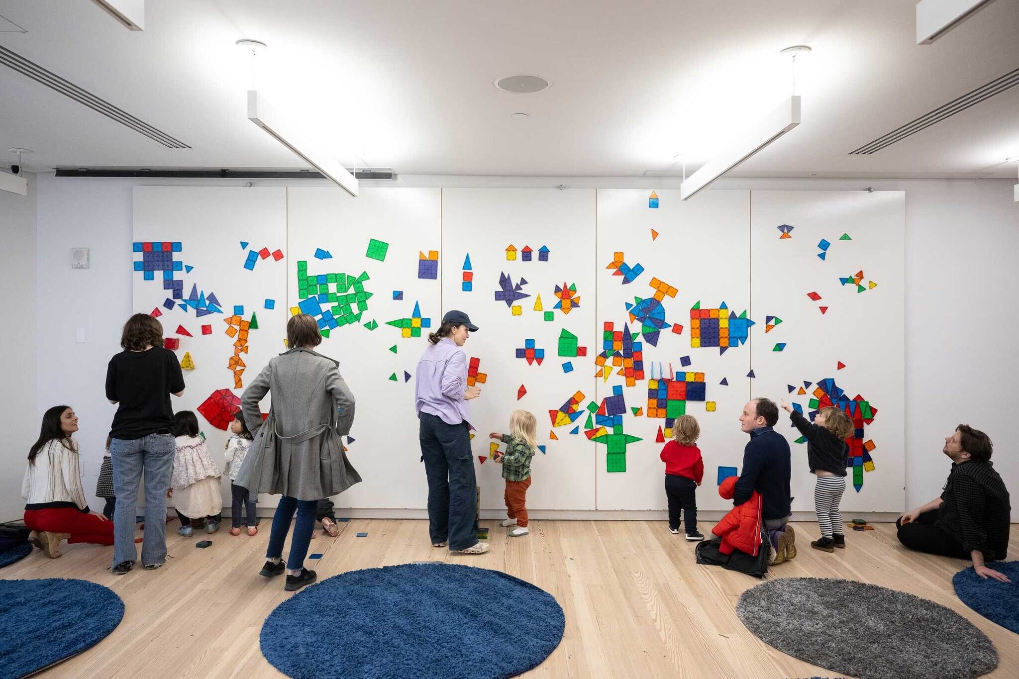 A group of adults and children are gathered in a brightly lit room at the Whitney Museum, engaging in a creative activity with colorful magnetic geometric shapes. The shapes are arranged on a large white wall, forming a vibrant mosaic. Some participants are standing, while others are kneeling or sitting on the floor. The room has a clean, modern design with round blue and gray rugs scattered on the wooden floor. The atmosphere appears lively and collaborative.