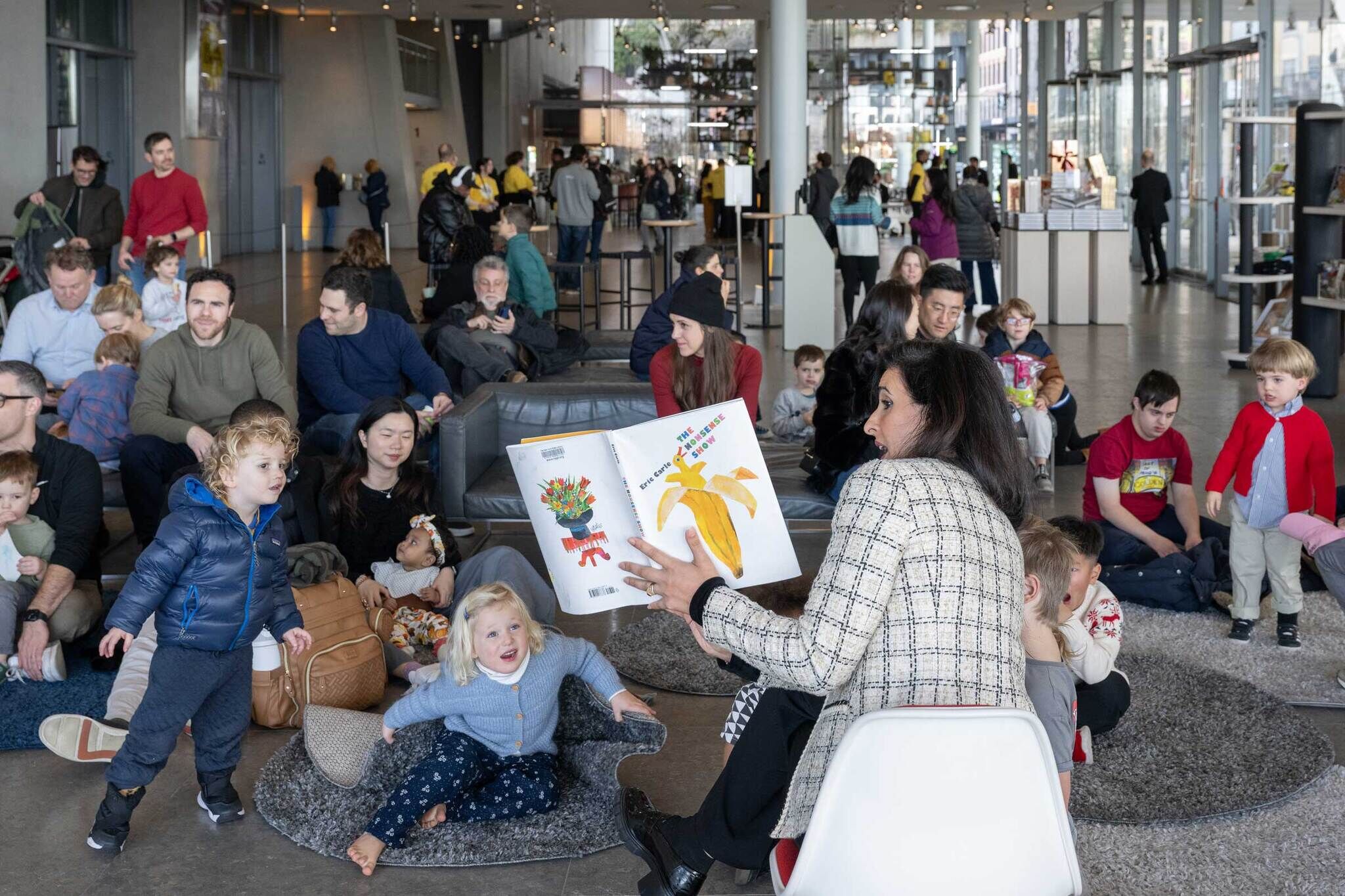 A woman reads a picture book to a group of children and adults in a busy, modern indoor space.