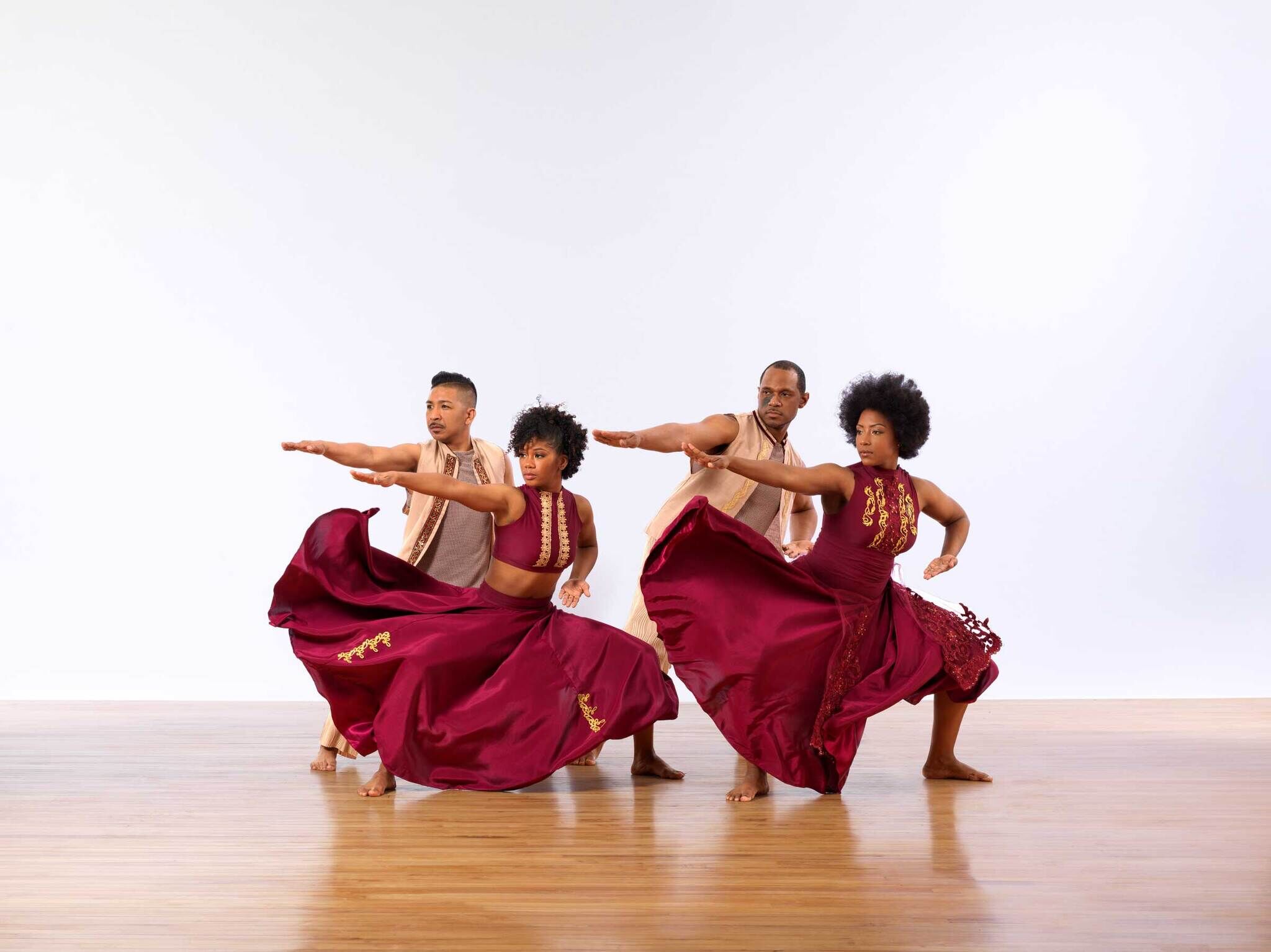 Four dancers in maroon outfits perform a synchronized pose with outstretched arms and flowing skirts on a wooden floor.