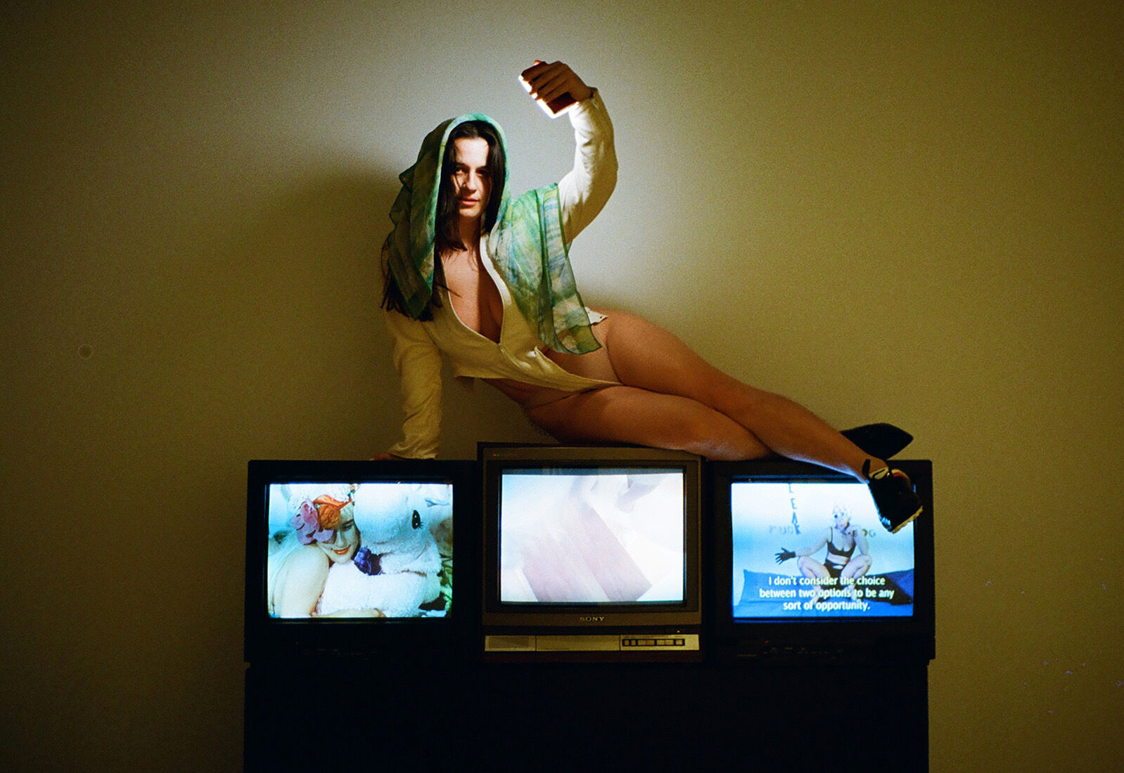 A woman in a partially unbuttoned blouse and green scarf, holding a smartphone up high with the screen shining onto her face, poses atop three CRT televisions displaying varied images in a dimly lit room.
