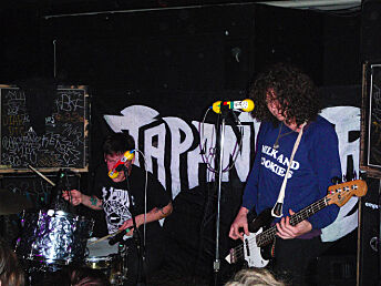 Two men playing music in front of a wall with Japanther painted on it.