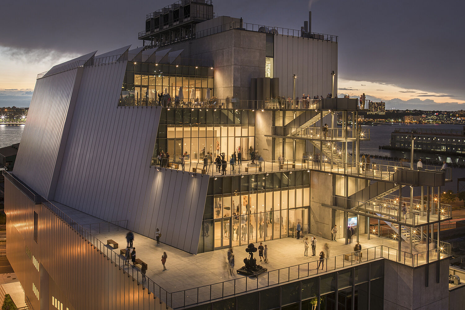 Exterior view of the Whitney Museum building at nighttime. The windows are brightly lit and museum-goers are visible in the galleries and on the terrace