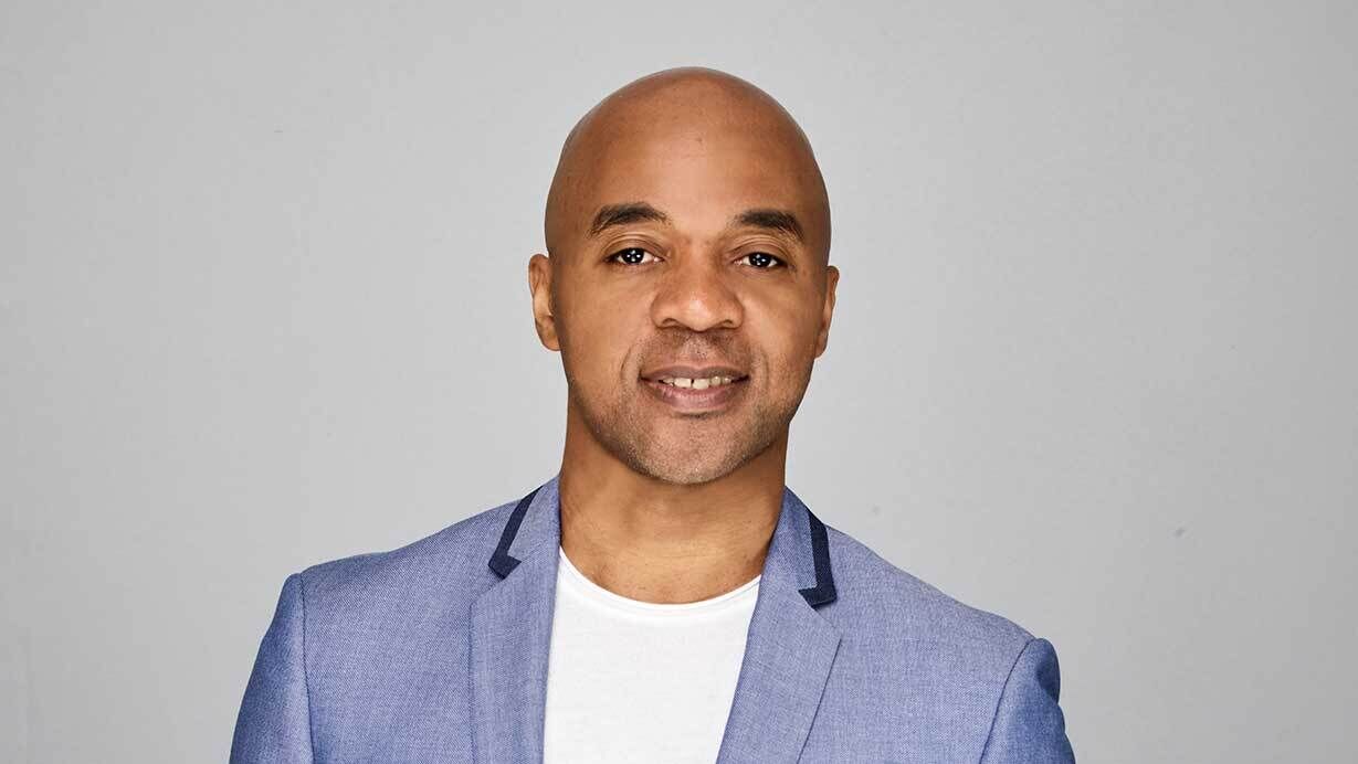 Man in a light blue blazer and white shirt, smiling against a plain gray background.