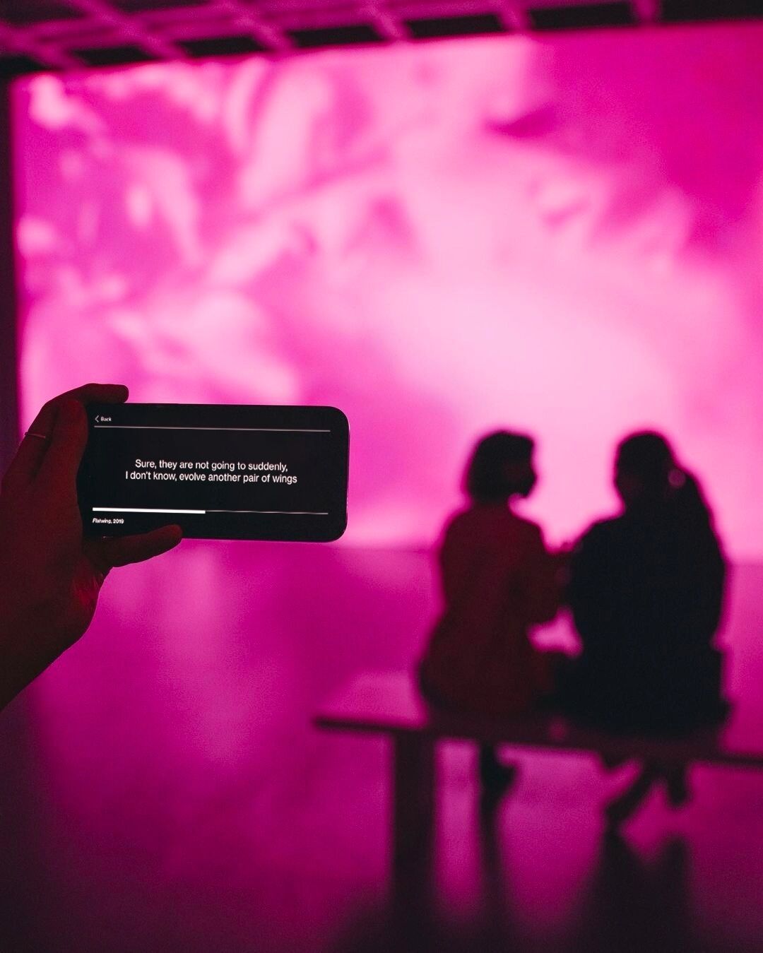 In the foreground, a hand holds a mobile device displaying white words on a black background and in the background, a two people view a fuchsia-soaked projection on a wall.