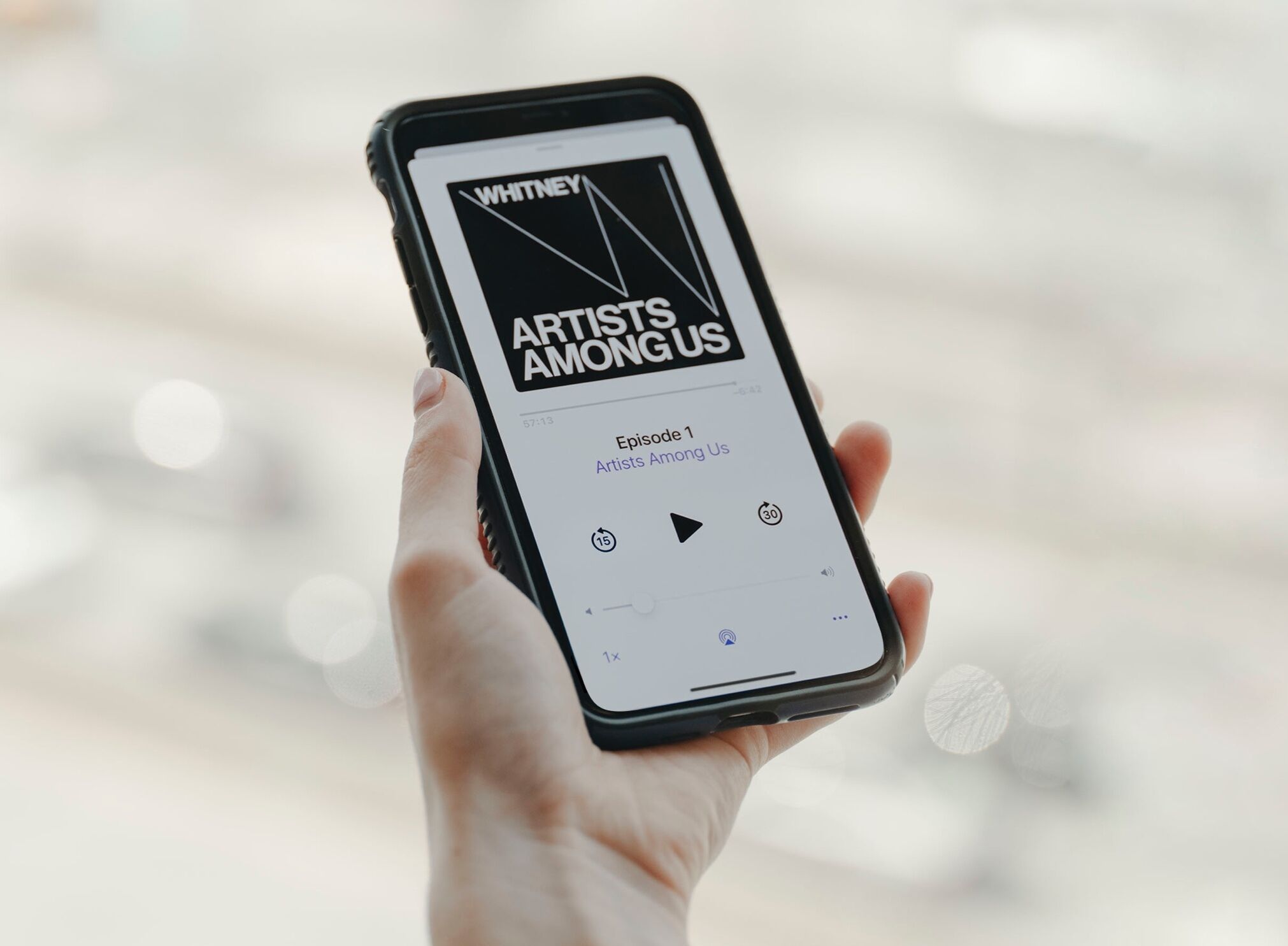 A hand holding a smartphone whose screen displays the Artists Among Us podcast playing.