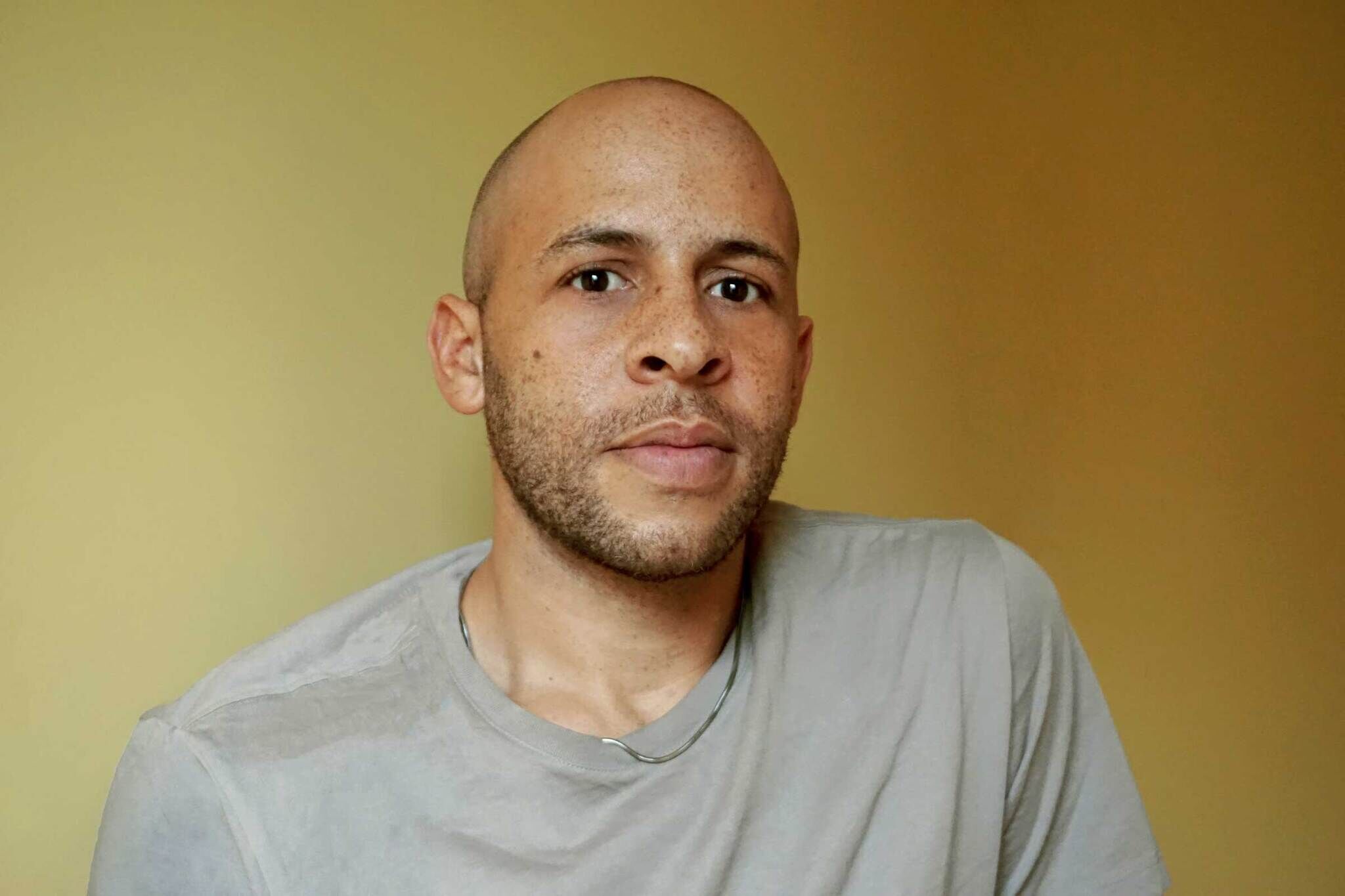 A man with a shaved head and light beard wearing a gray t-shirt, looking directly at the camera against a plain yellow background.