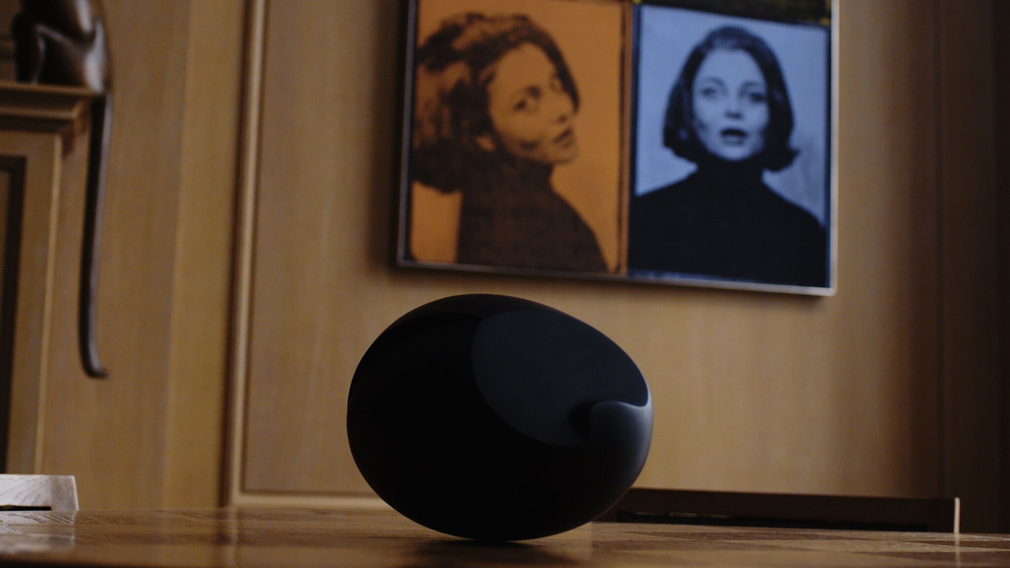 A video still of a round object on the floor in front of a wall with portraits hanging on it.