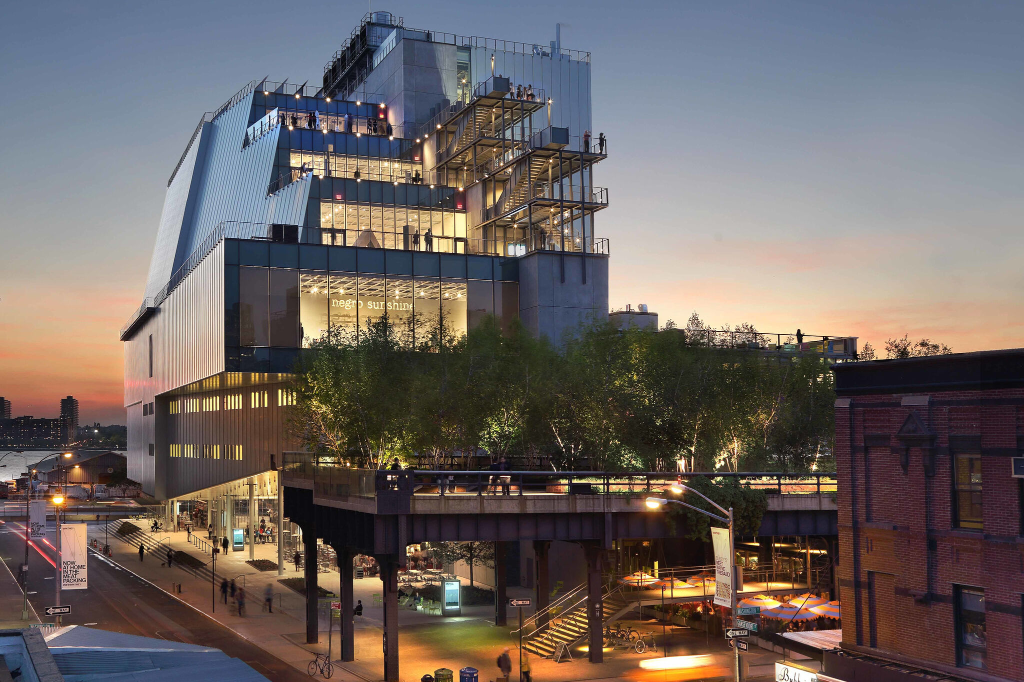 Modern building with external staircases at dusk, overlooking an urban street with pedestrians and cars.