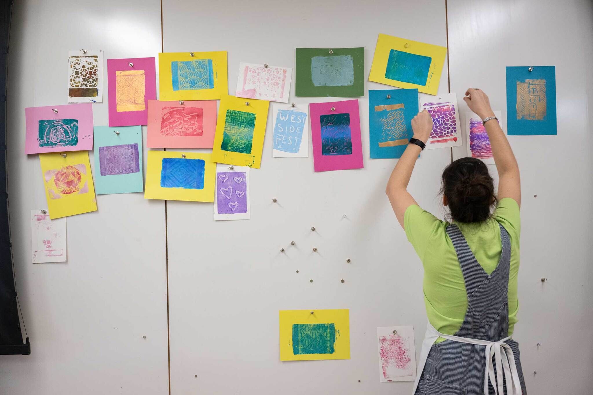 Person in a green shirt and apron pins colorful artwork to a white wall, with various prints and patterns already displayed.