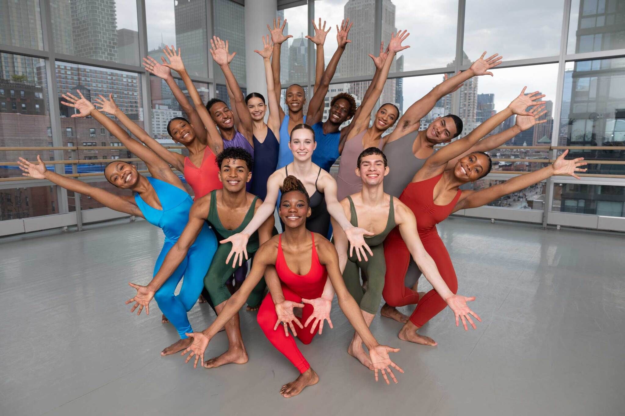 A group of dancers in colorful outfits pose energetically with arms raised in a dance studio with large windows and a cityscape in the background.