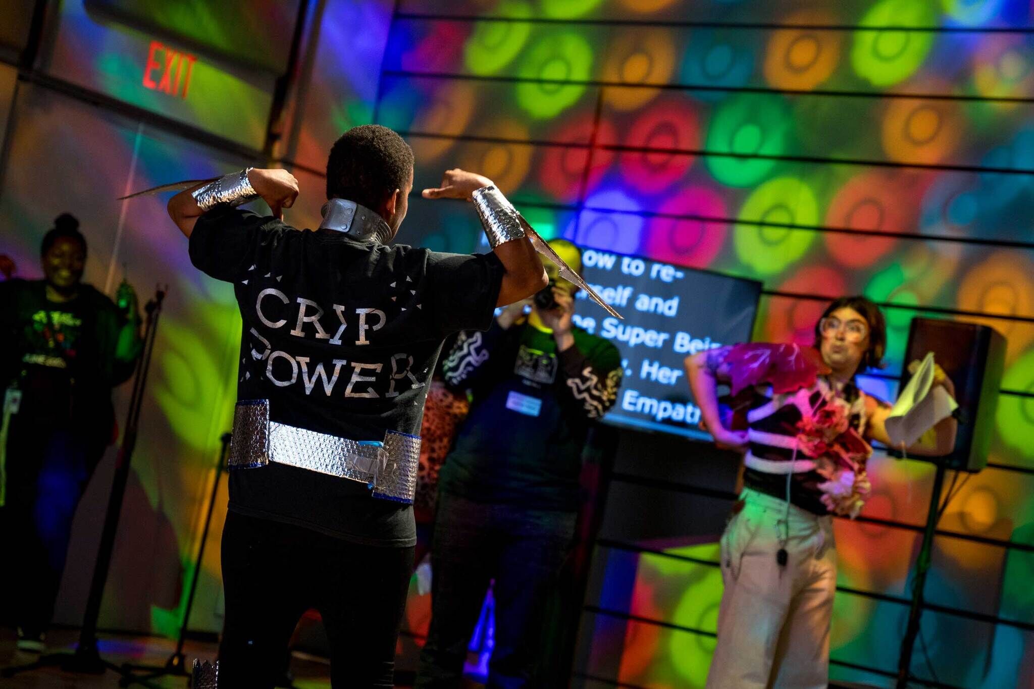 Radical Joy Ball participant poses and flexes their muscles while wearing a shirt that says Crip Power on the back