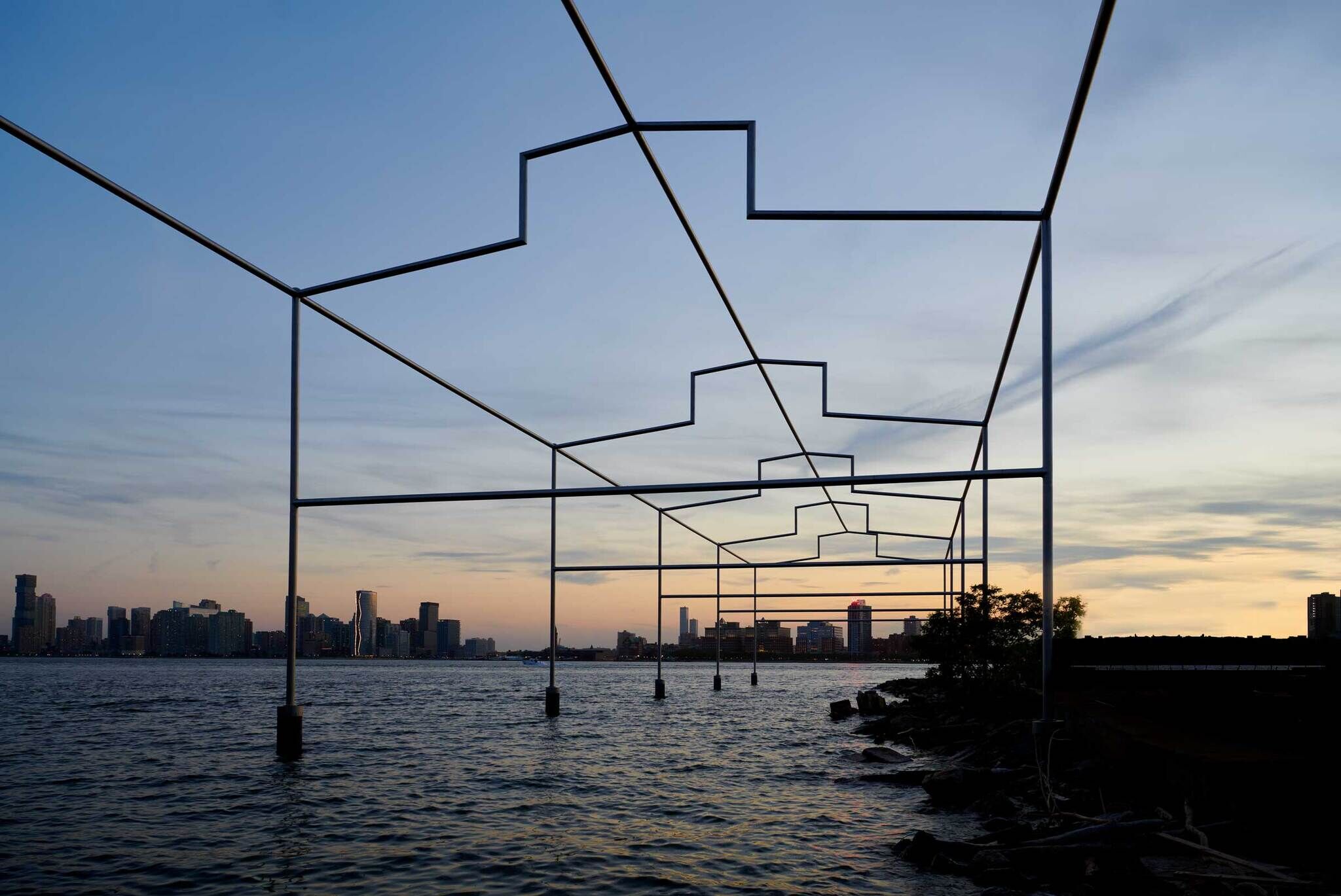 A metal art sculpture over water with a city skyline in the background at sunset.