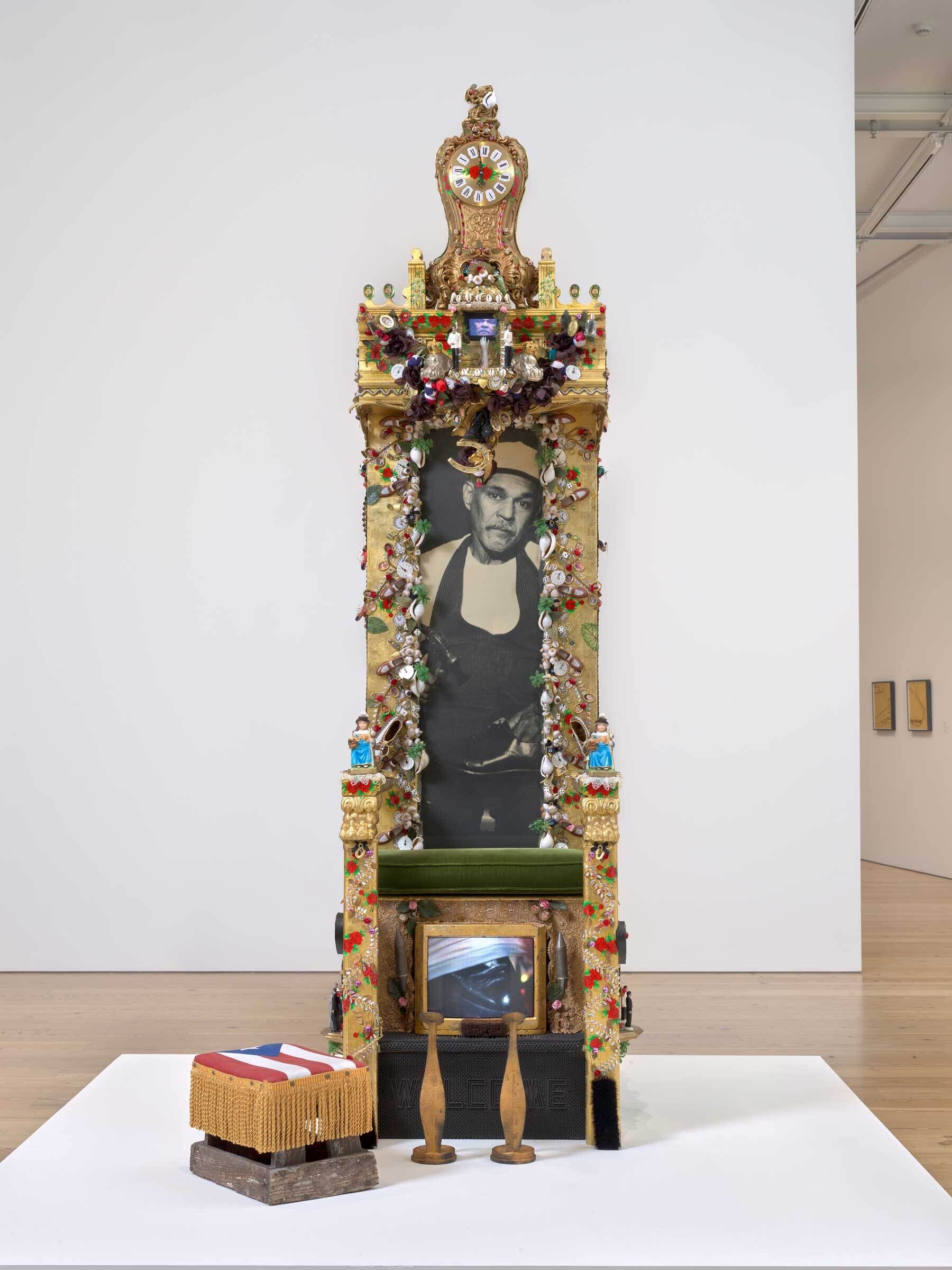 A mixed-media artwork resembling a throne, with a black-and-white portrait of a man as the central focus. The throne is adorned with intricate gold details, colorful jewels, and various found objects, creating an ornate and shrine-like appearance. Below the photograph is a small screen or reflective surface, adding a dynamic element. In front of the throne is a low stool covered in fabric featuring an American flag design, along with two wooden pins resembling exercise clubs. 