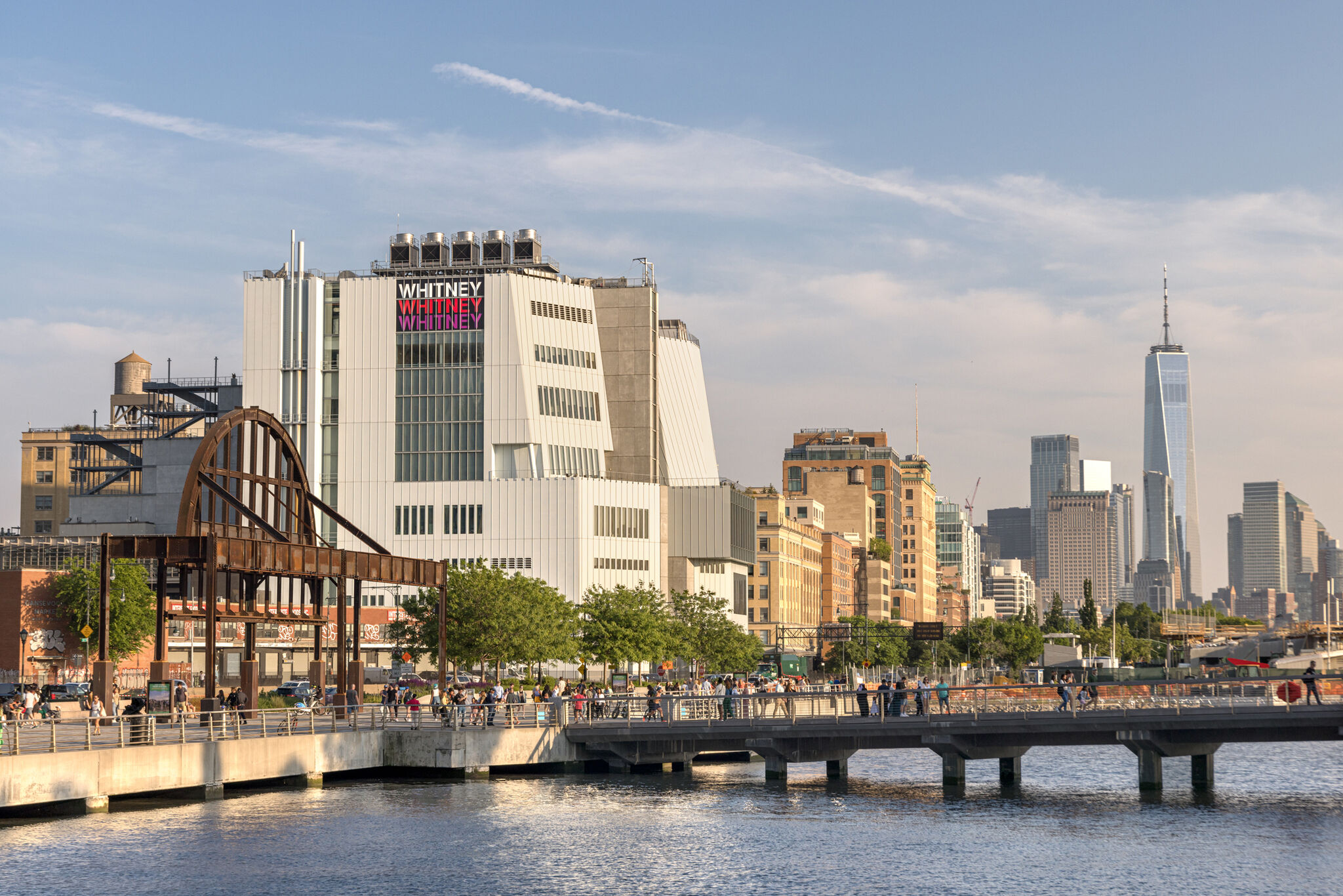 The Whitney Museum in NYC with the One World Trade Center in the background, viewed from a riverside walkway on a sunny day.