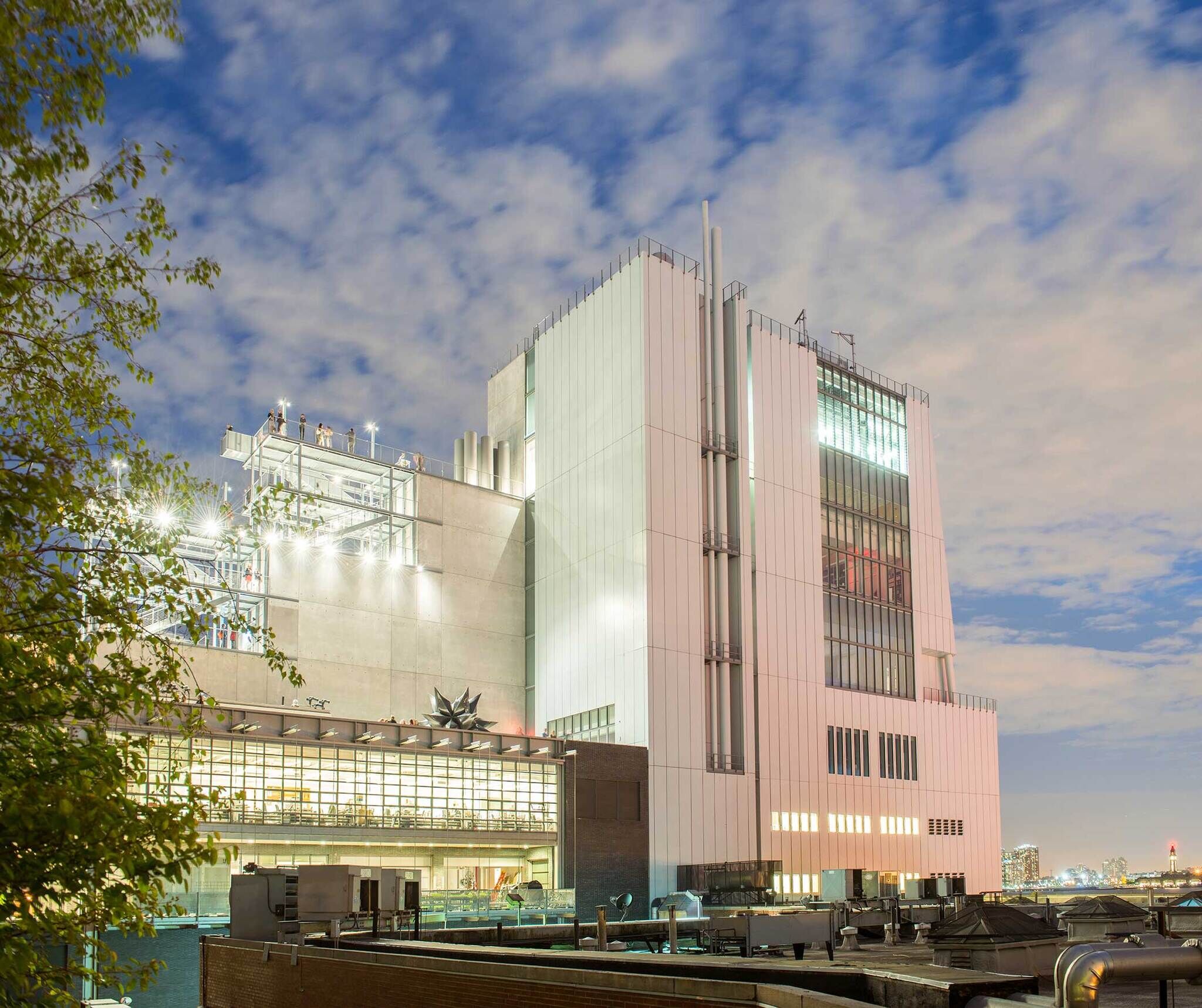 Modern building with large windows and rooftop terrace, set against a partly cloudy sky. Trees and cityscape visible in the background.