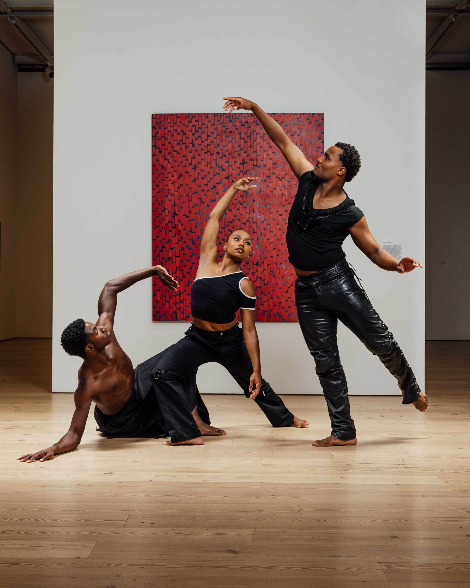 Three Black dancers, a woman flanked by two men, pose with dramatic and graceful dance gestures in a gallery in front of an abstract painting with thick vertical red brushstrokes running from edge to edge with a shimmering dark background peeking through.