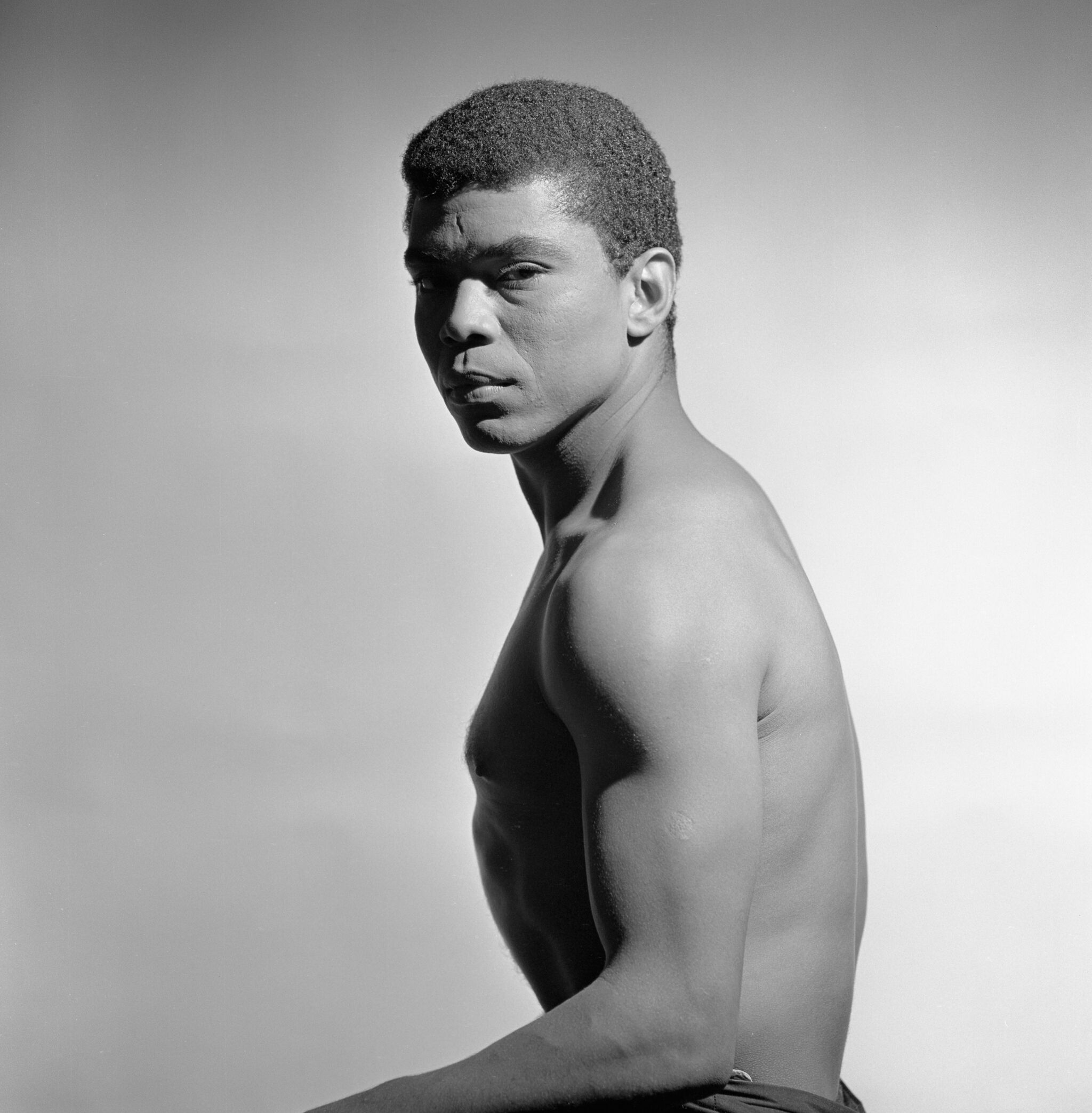 Black and white photo of a shirtless man with short hair, looking over his shoulder with a serious expression against a plain background.