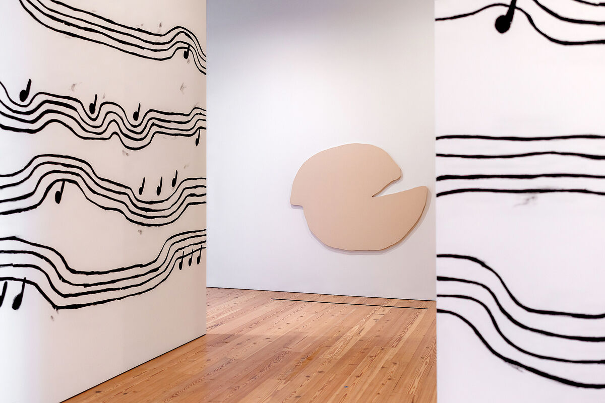 Gallery with abstract art; musical notes on wavy lines on walls, beige irregular shape on another wall, wooden floor.